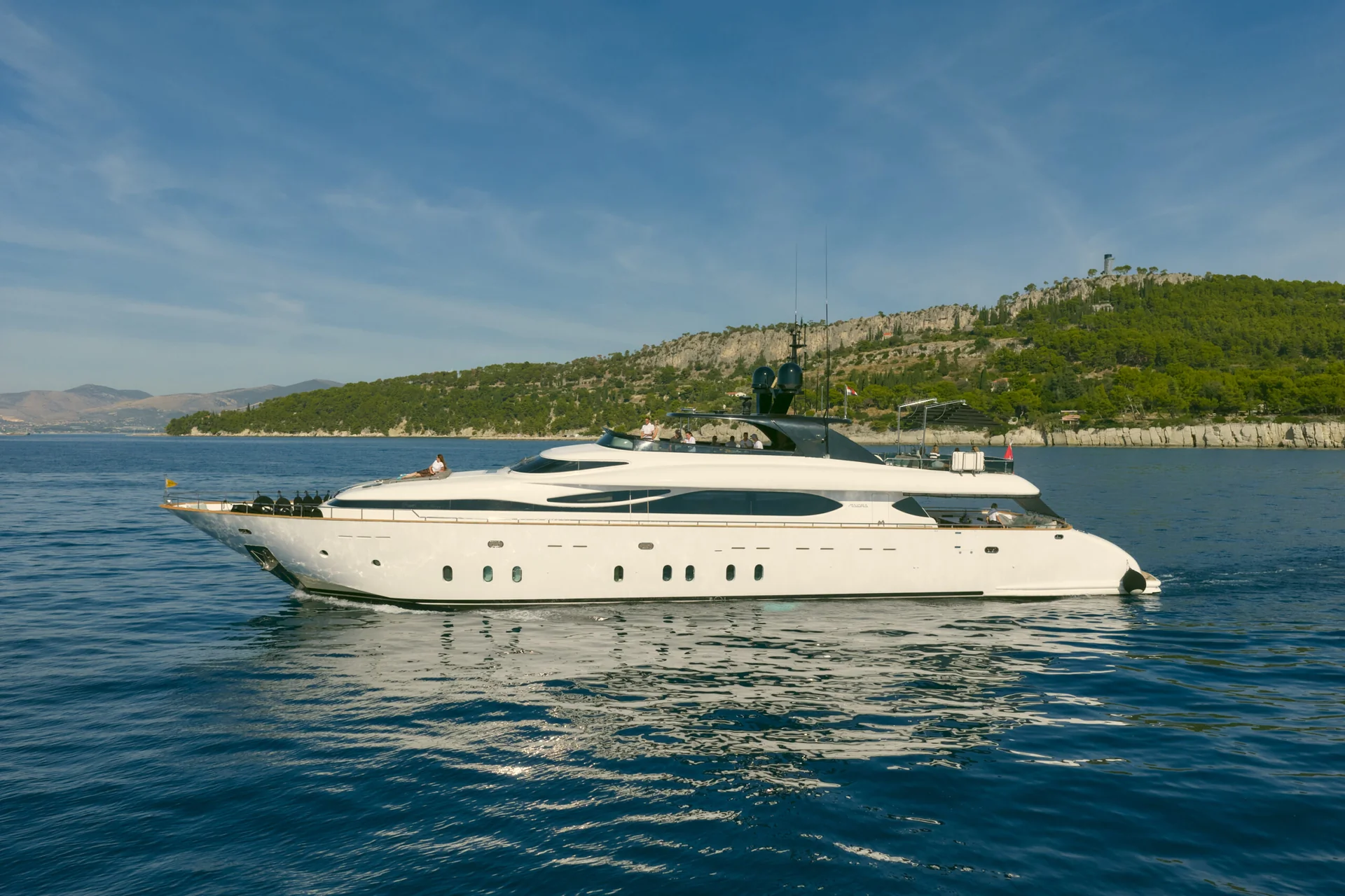A white luxury yacht cruising on calm blue water with a forested coastline in the background.
