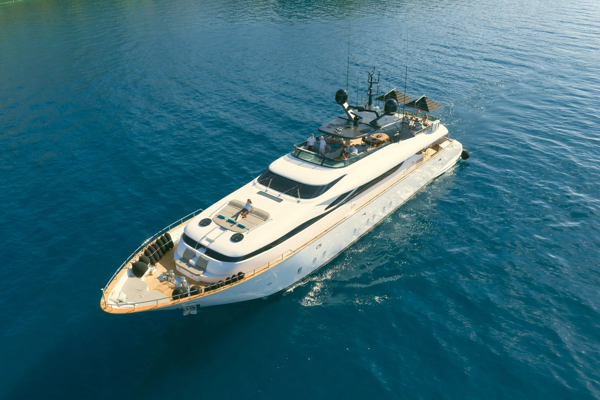 Aerial view of a white yacht cruising on deep blue water, with several people lounging on the decks.
