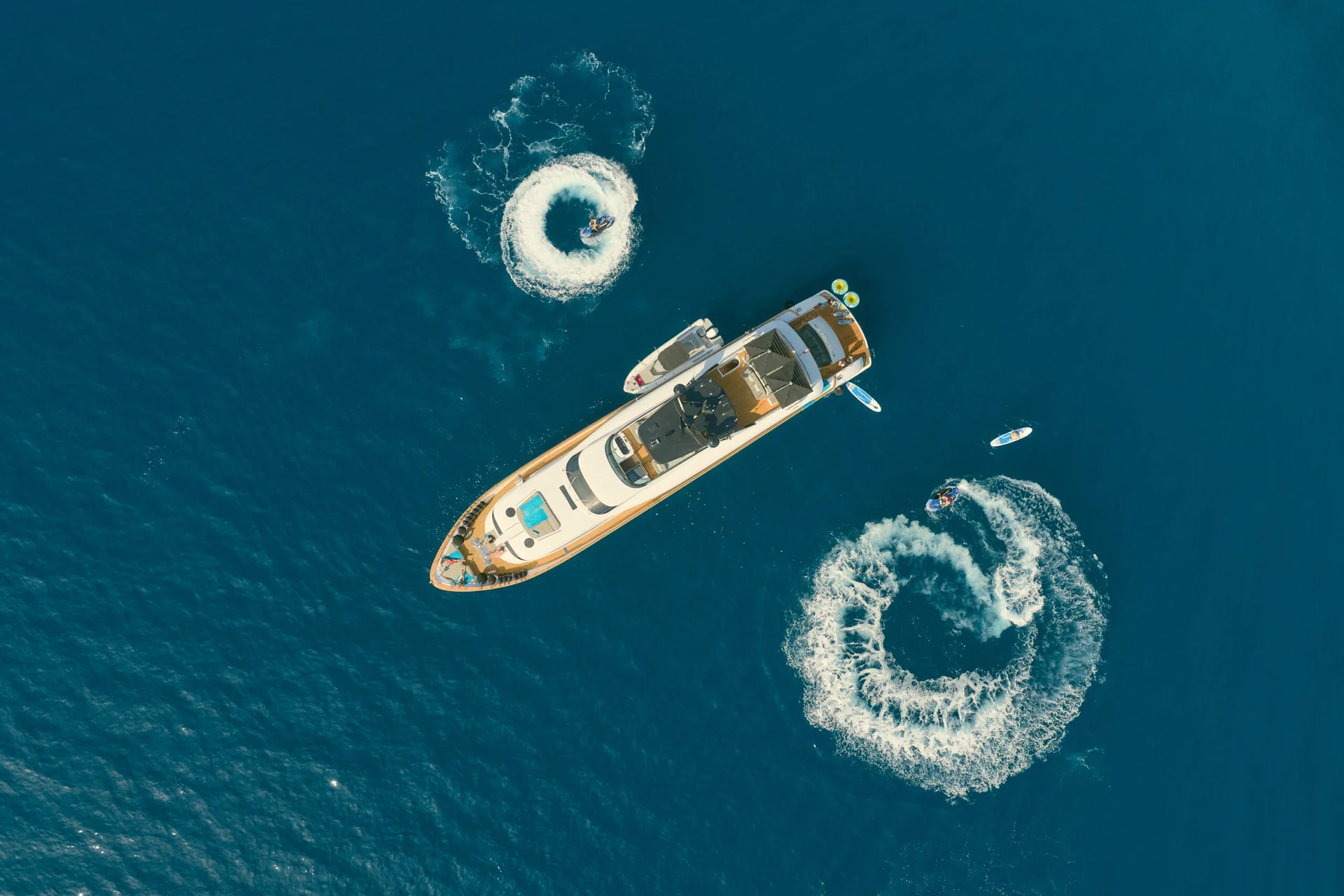 Aerial view of a yacht in the open sea, surrounded by two jet skis creating circular wakes and a person on a paddleboard.