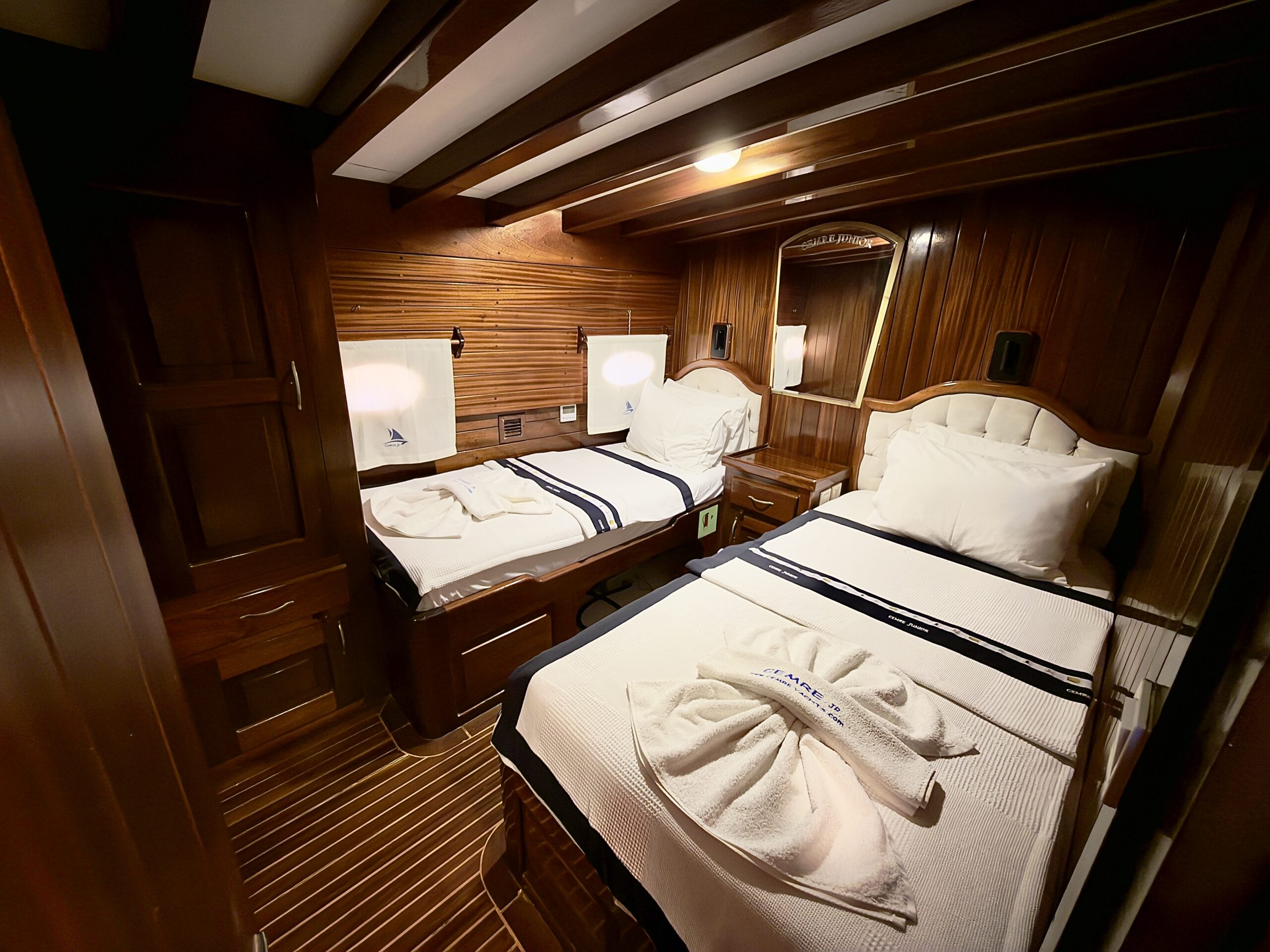 CEMRE JUNIOR A cozy cabin bedroom with two single beds, wooden walls and ceiling, white bedding, reading lamps, and a towel art decoration on one bed. Soft lighting creates a warm, inviting atmosphere.