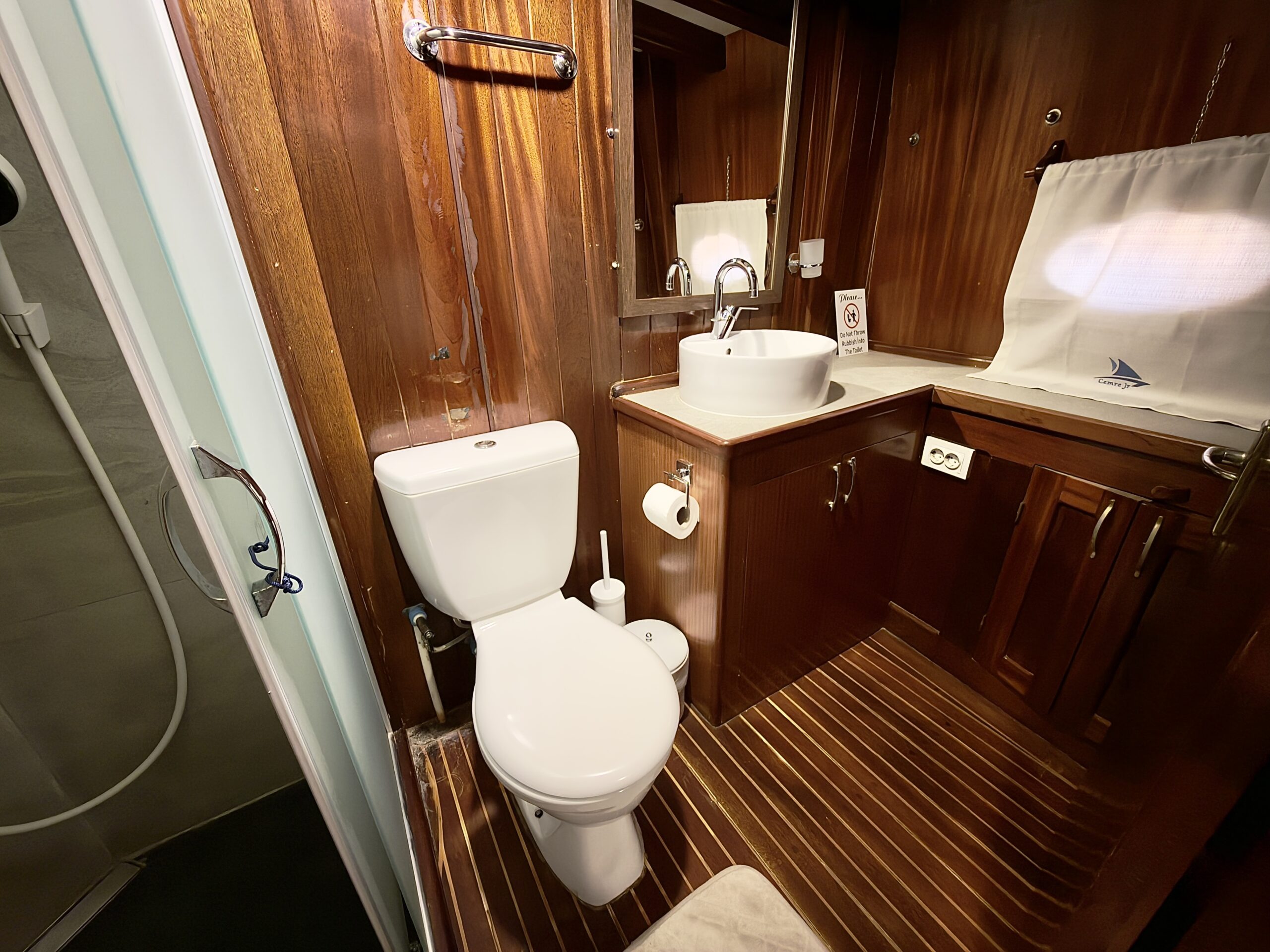 CEMRE JUNIOR A small wooden bathroom with a toilet, sink, mirror, cabinet, and shower area. There is a toilet brush, toilet paper, towel rack, and a curtain covering a window. The floor and walls are wood-paneled.