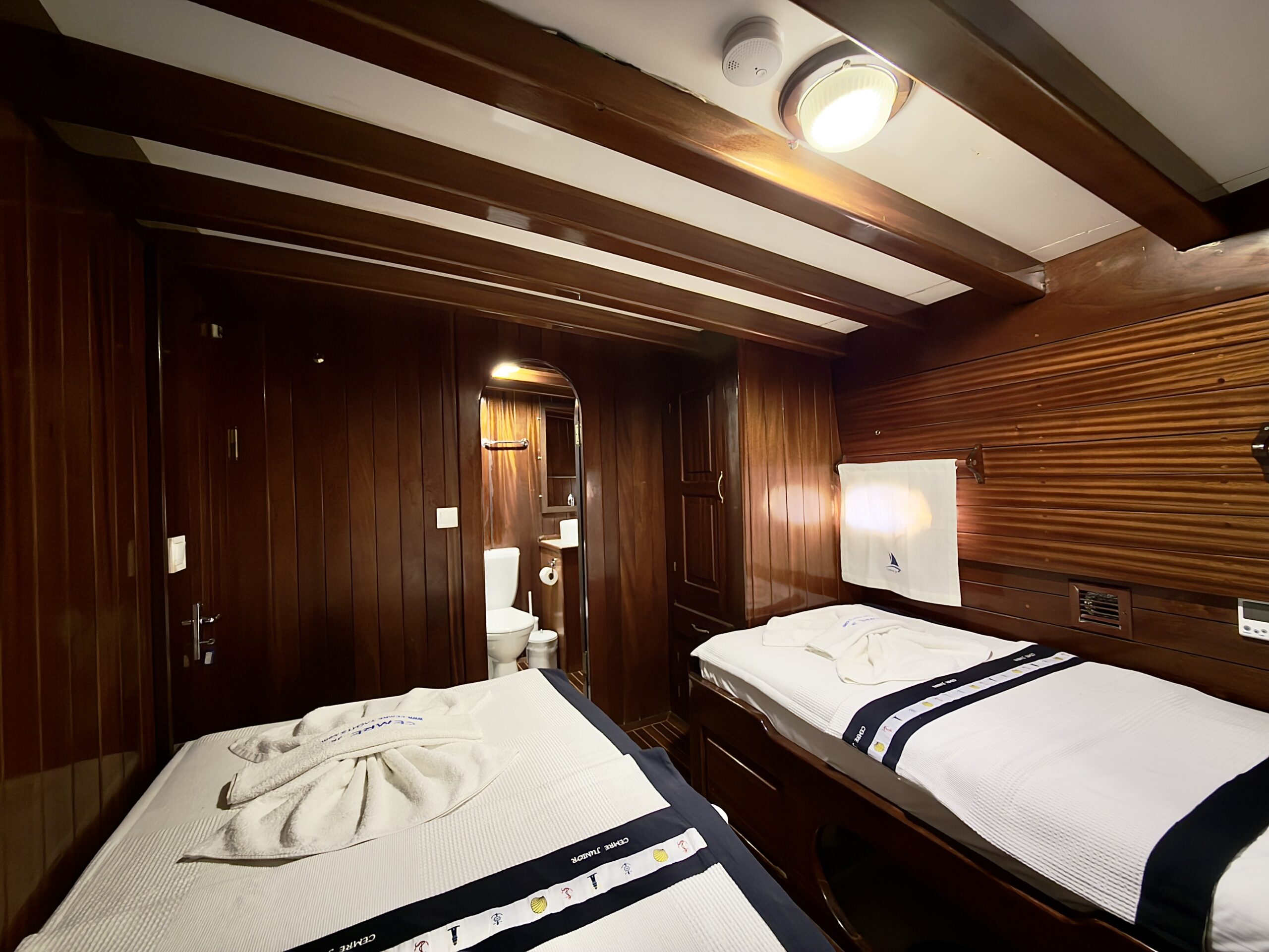 CEMRE JUNIOR A cozy wooden cabin with two single beds, neatly made with white linens and towels. The room has wood-paneled walls and ceiling, and an en-suite bathroom visible in the background. Warm overhead lighting brightens the space.