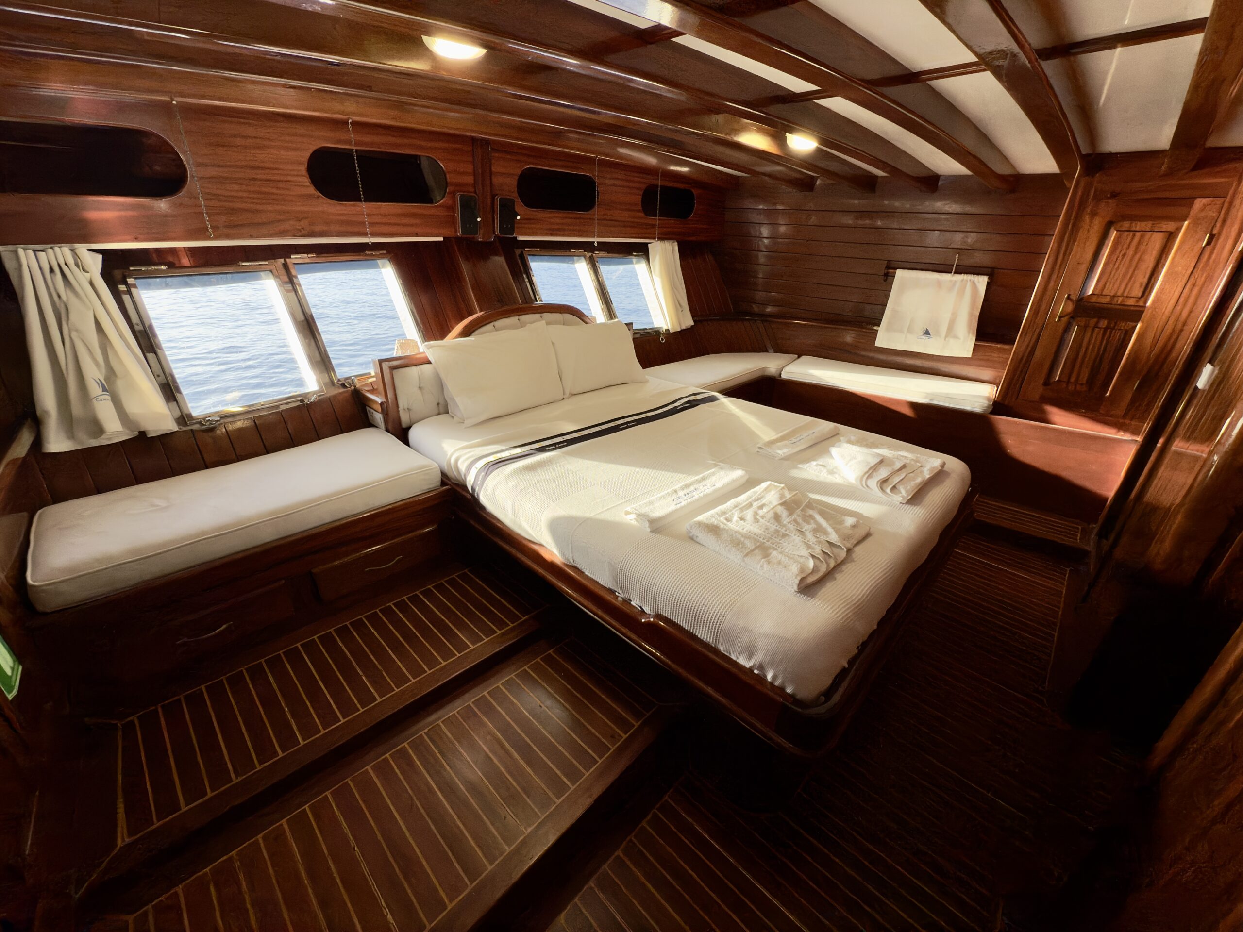 CEMRE JUNIOR A cozy wooden cabin on a boat with a large bed, white linens, two folded towels, windows overlooking the sea, and built-in benches along the walls. Warm lighting enhances the polished wood interior.