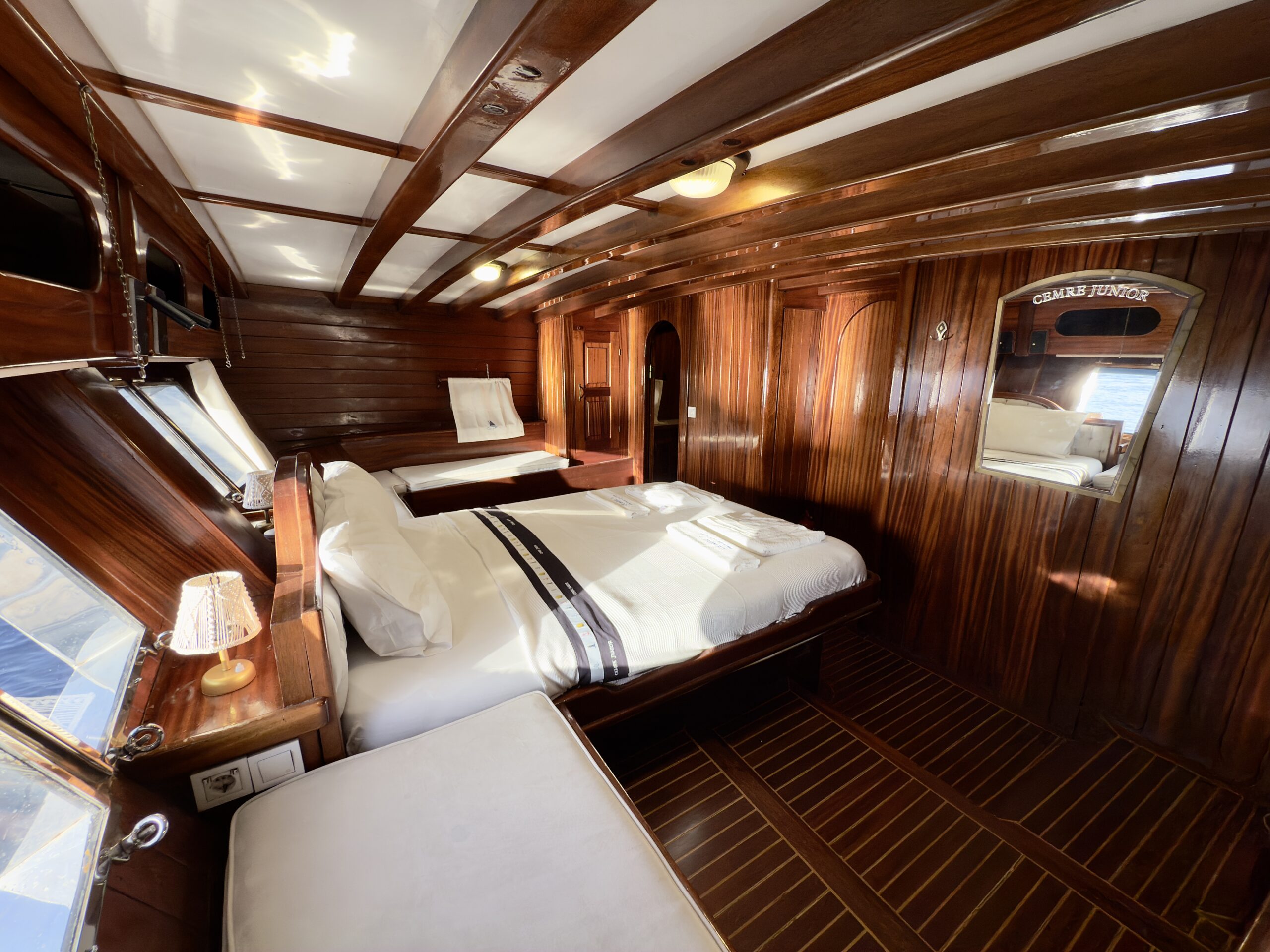 CEMRE JUNIOR Luxurious wooden yacht cabin with polished dark wood walls and ceiling, a large bed with white linens, a mirror, and windows letting in sunlight. A small lamp and folded towels rest on the bedside table.