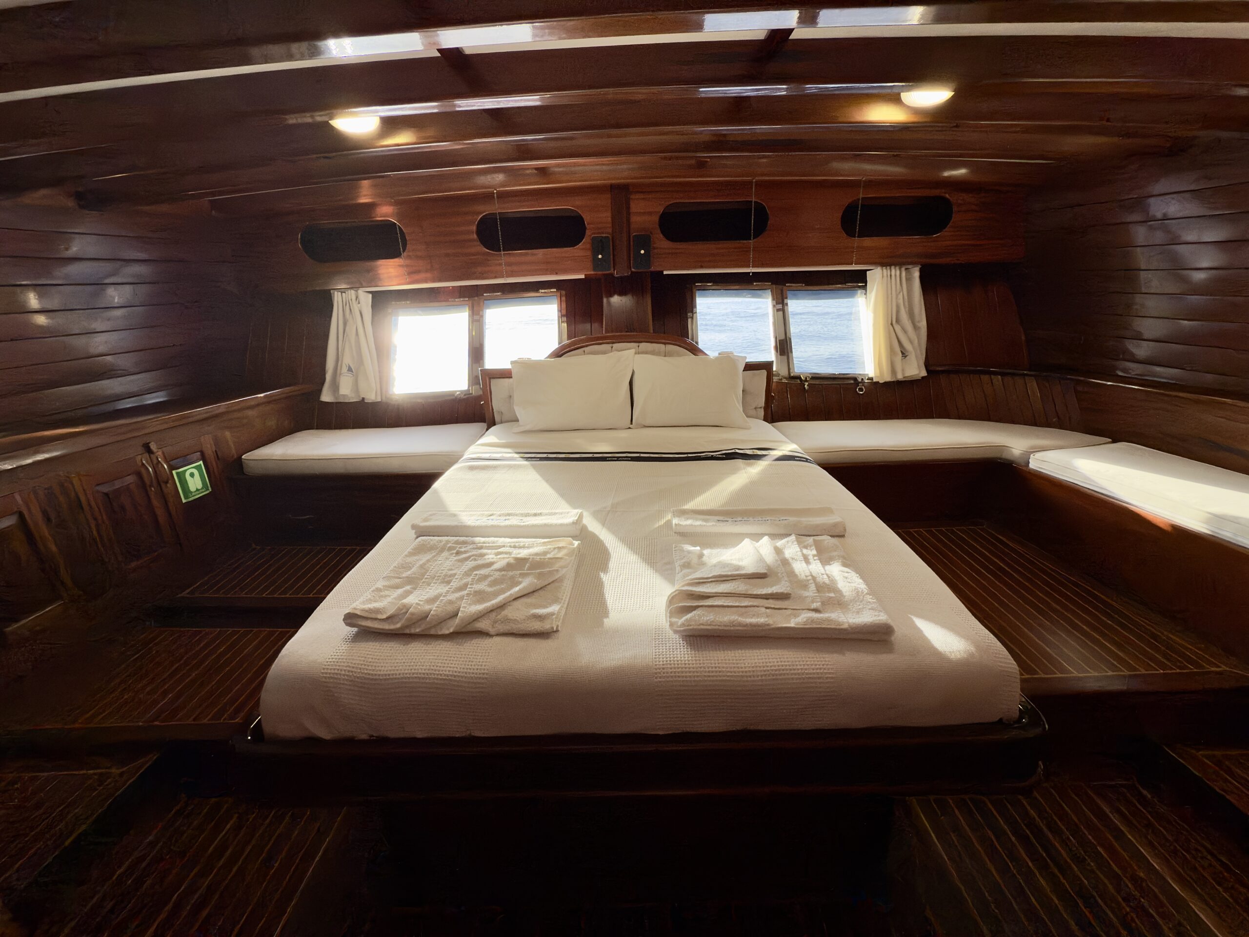 CEMRE JUNIOR A cozy wooden boat cabin with a neatly made double bed, two white towels, soft lighting, and windows at the end of the bed showing a view of the sea.