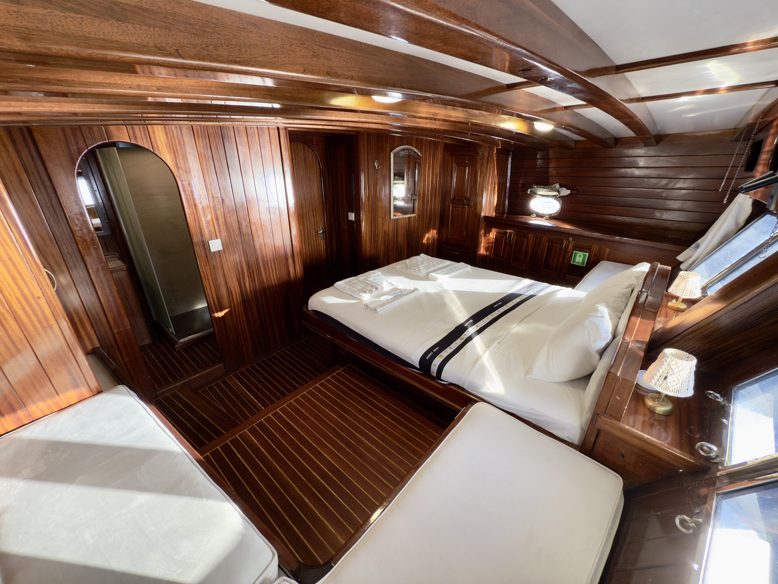 CEMRE JUNIOR A cozy wooden cabin with polished paneling, a double bed neatly made with white linens, a narrow daybed, built-in mirrors, soft lighting, and windows letting in natural sunlight.