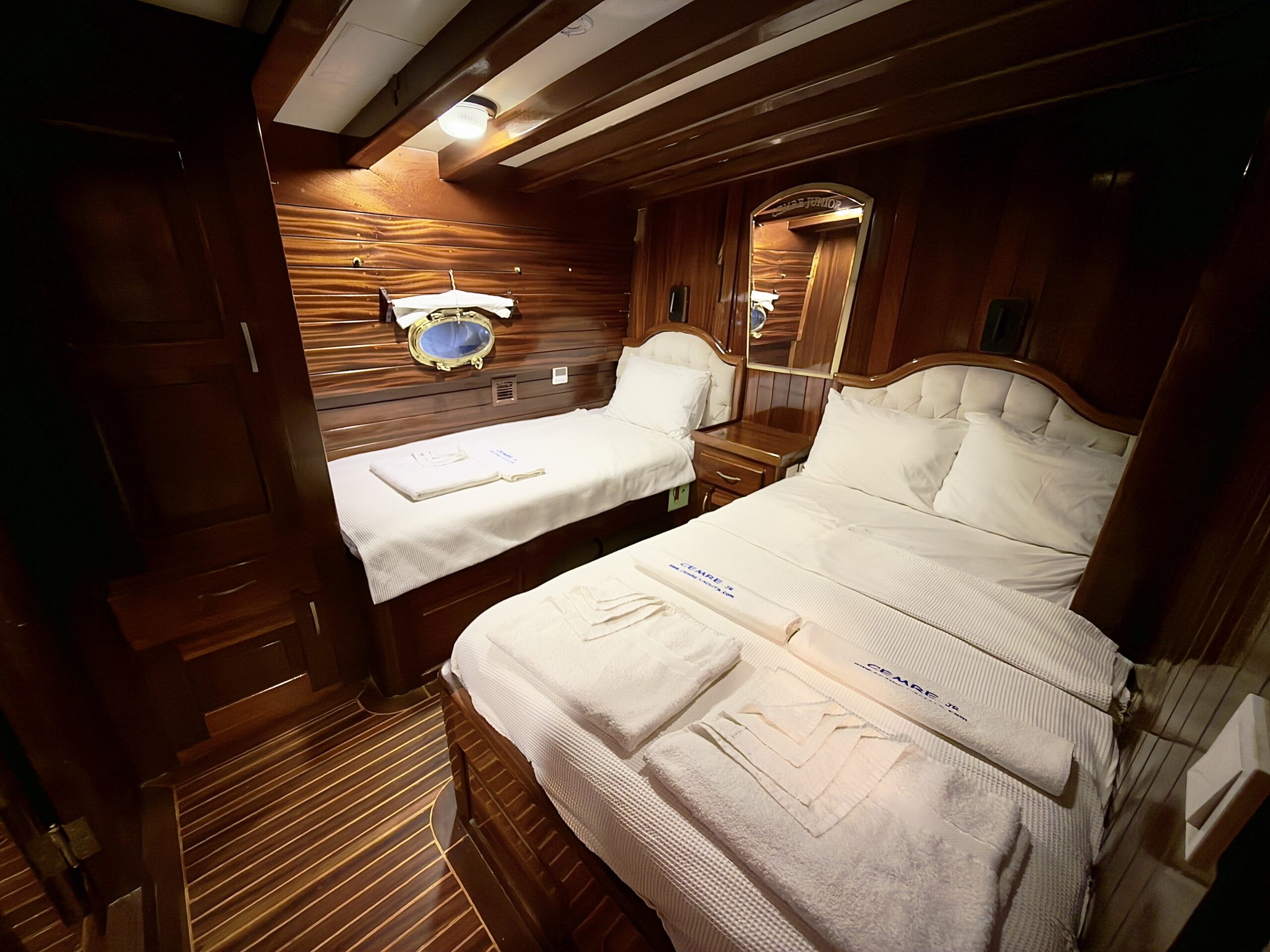 CEMRE JUNIOR A cozy wooden cabin bedroom with a double bed and a single bed, both neatly made with white linens and towels. There is a small window with a nautical frame, a mirror, and warm lighting creating an inviting atmosphere.