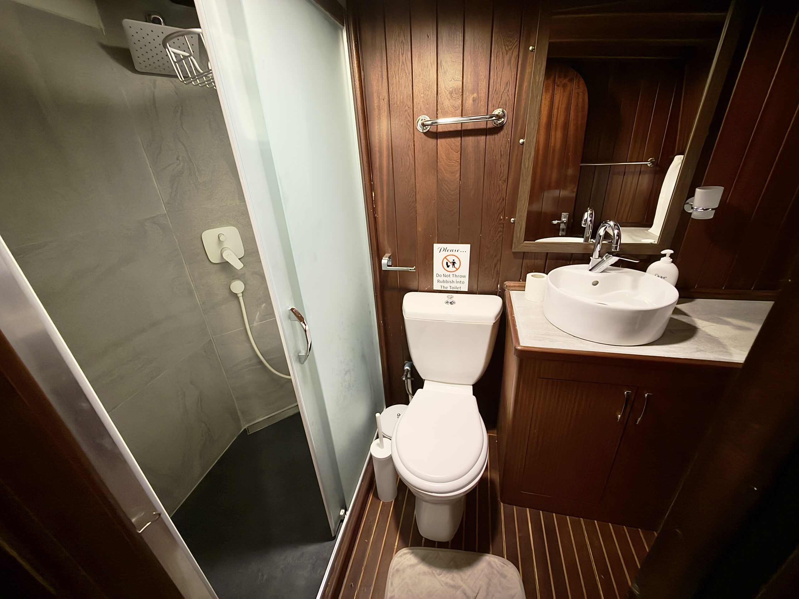 CEMRE JUNIOR A compact bathroom with a wooden interior, including a toilet, white sink on a vanity, mirror, and a glass-enclosed shower with a handheld showerhead. A small sign is placed on top of the toilet tank.