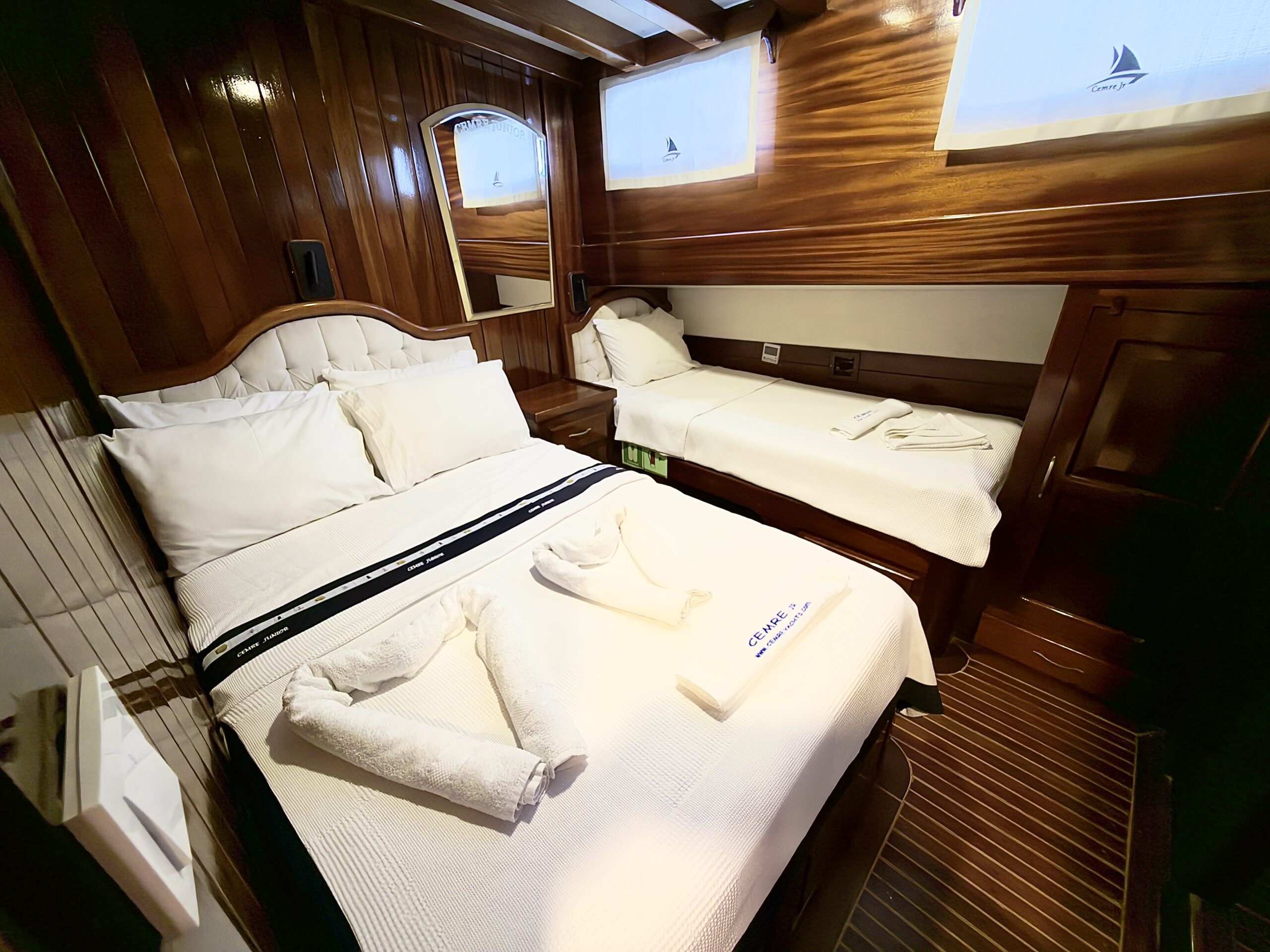 CEMRE JUNIOR A cozy cabin room with polished wooden walls, a double bed and a single bed, both neatly made with white linens. Towels are arranged on the double bed, and sunlight filters through small windows.