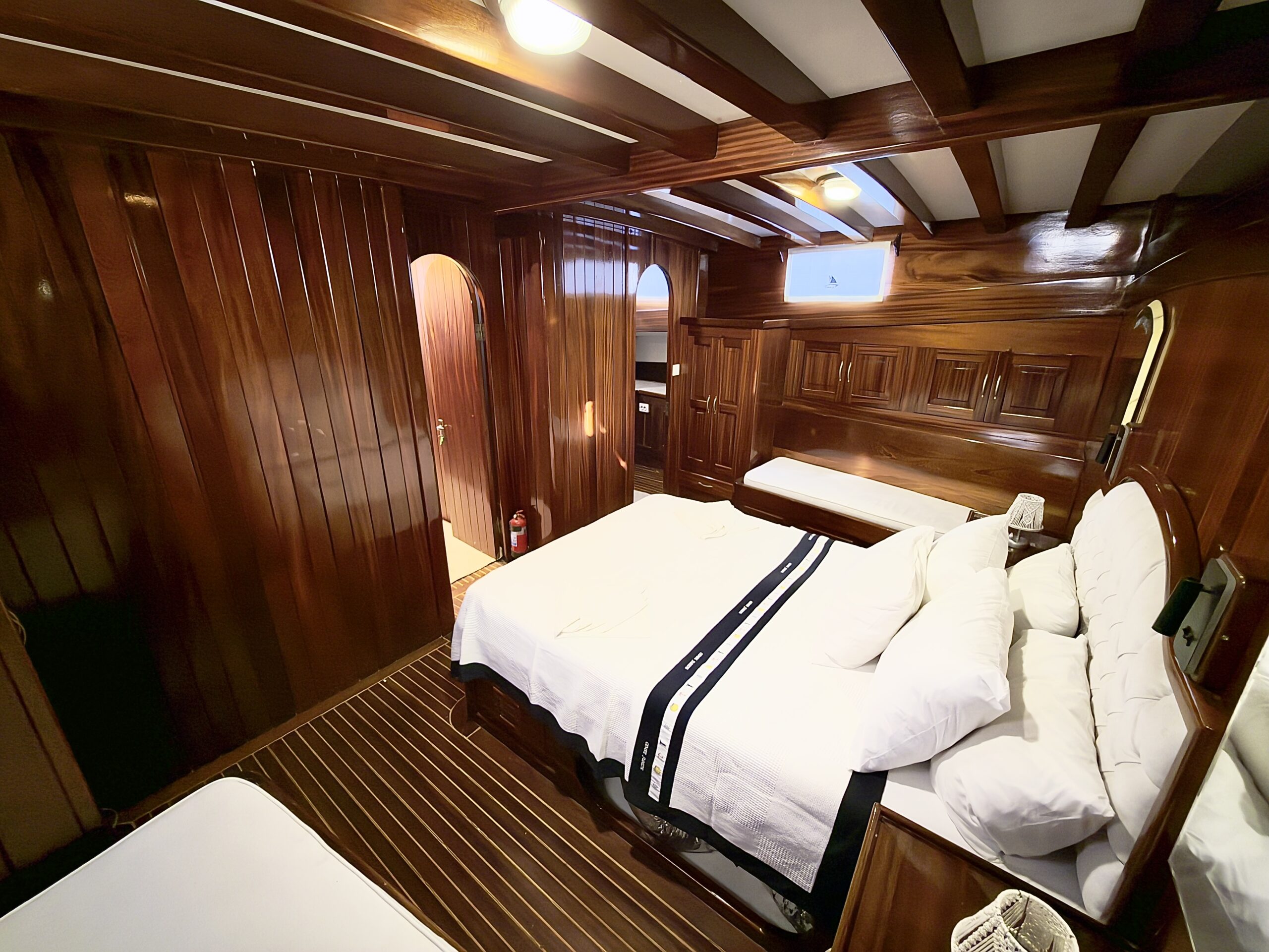 CEMRE JUNIOR A cozy bedroom with wooden walls, ceiling beams, and flooring, featuring a neatly made bed with white linens, a striped blanket, and a small window letting in light. Theres a door leading to another room.