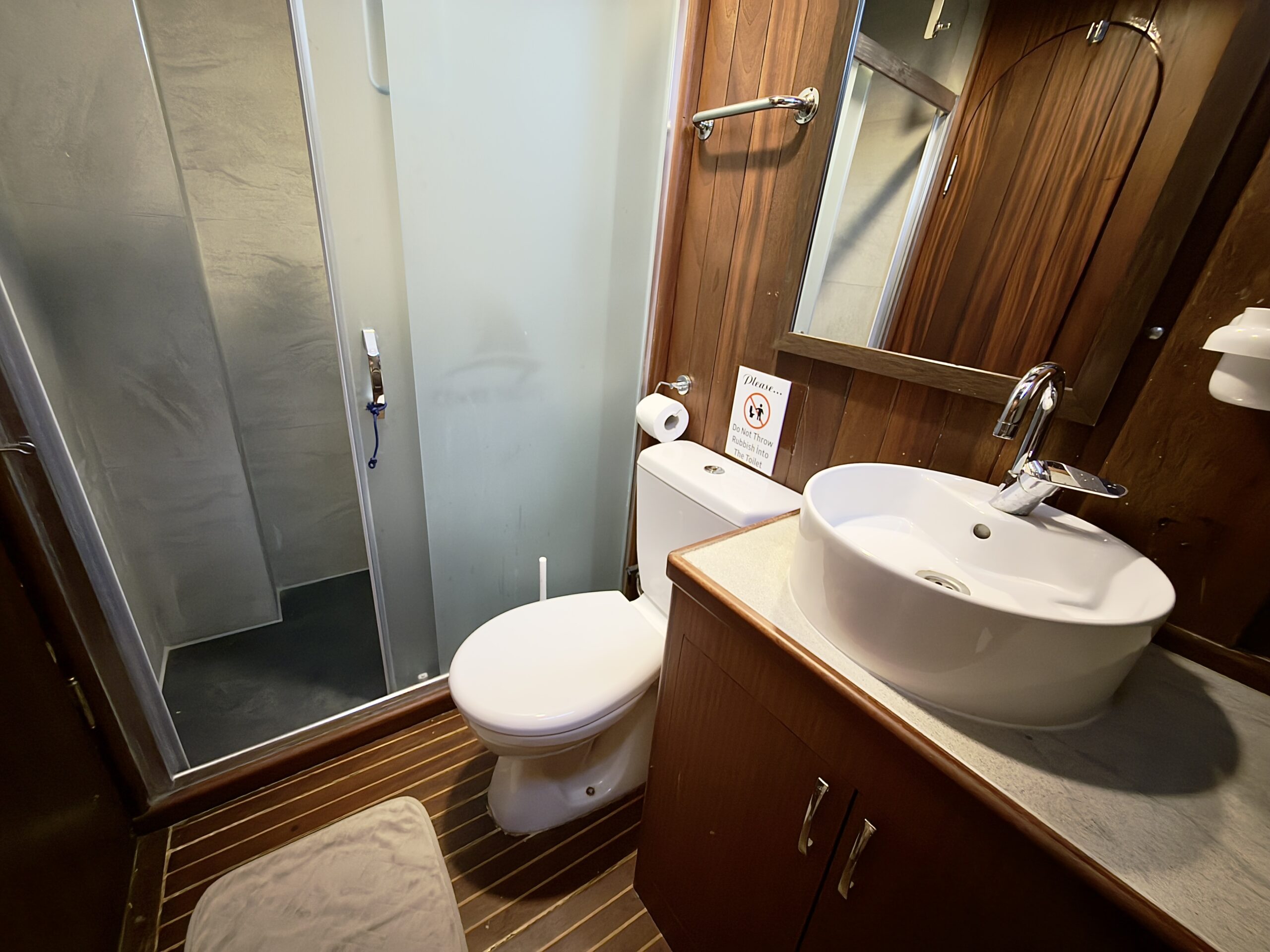 CEMRE JUNIOR A small bathroom with wood-paneled walls and floor, featuring a glass shower, white toilet, round vessel sink on a wooden vanity, mirror, towel holder, and a toilet paper holder.