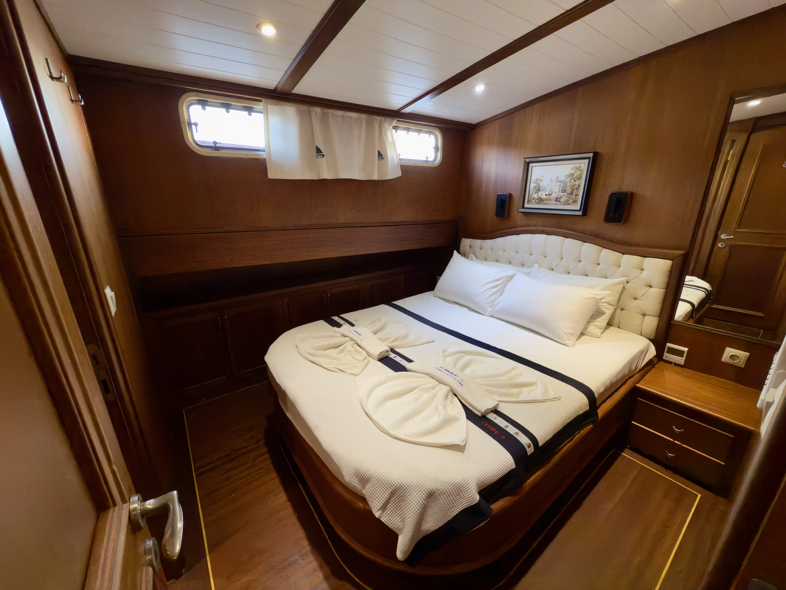 CEMRE 4 A cozy yacht cabin with wood-paneled walls features a large bed with white linens, towel art, a tufted headboard, porthole windows, a framed painting, and warm lighting.