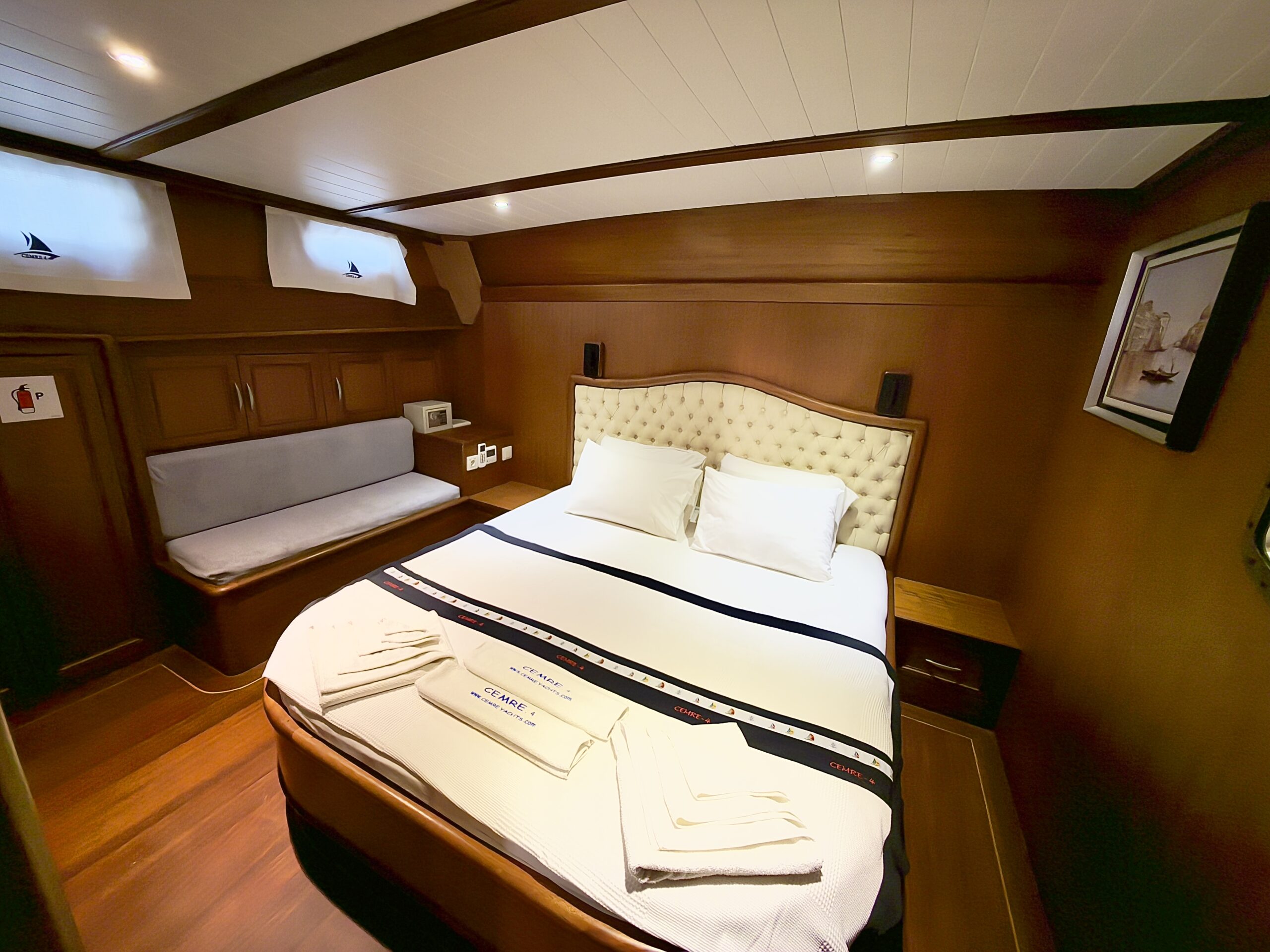 CEMRE 4 A cozy, wood-paneled bedroom with a double bed, white linens, folded towels at the foot, a cushioned bench by the window, built-in cabinets, and nautical-themed decor. Warm, ambient lighting creates an inviting atmosphere.