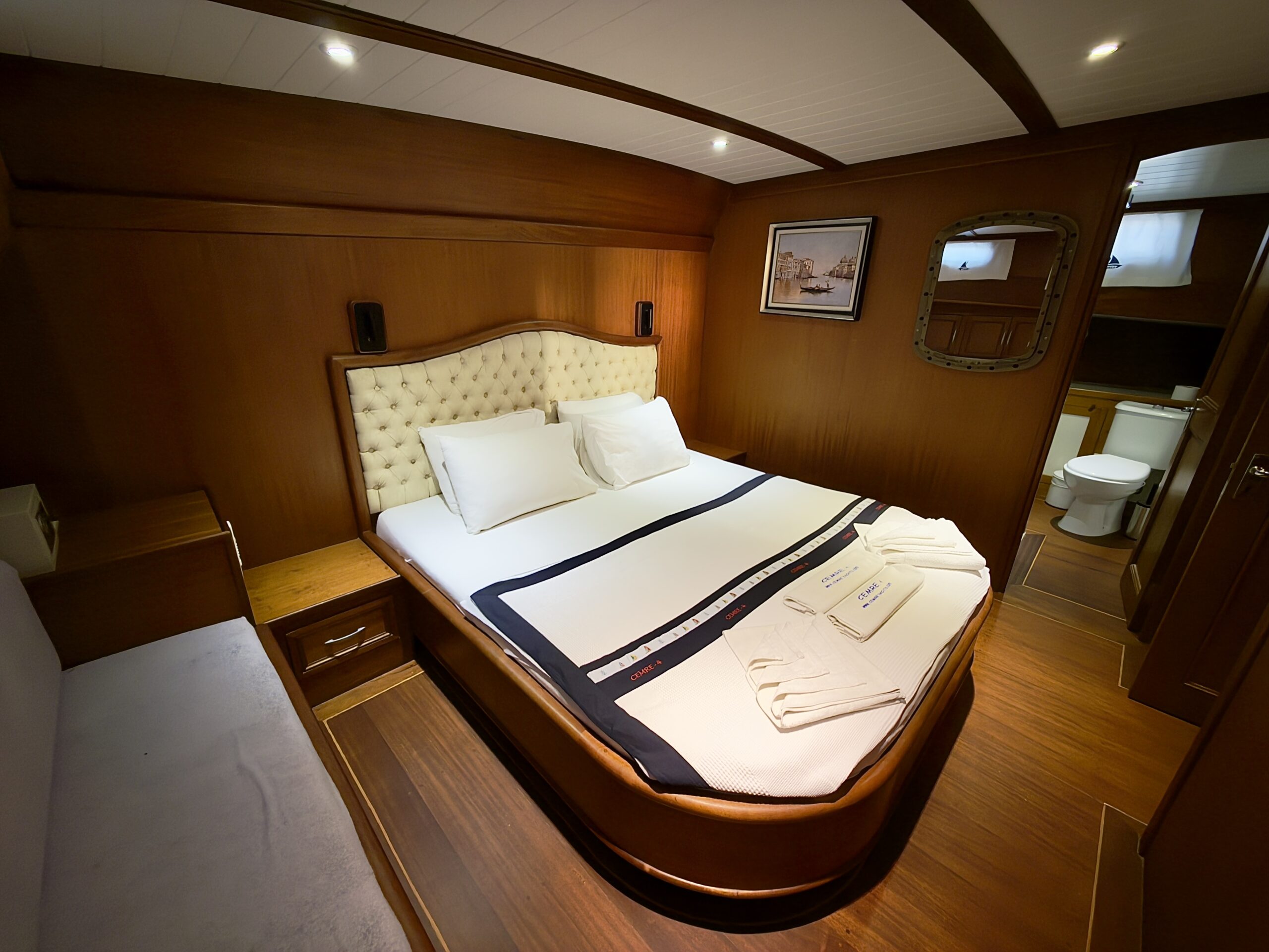 CEMRE 4 A cozy, wood-paneled bedroom with a double bed, tufted headboard, and white linens. Towels are neatly folded on the bed. Theres a small bathroom visible through an open door, and framed nautical art hangs on the wall.