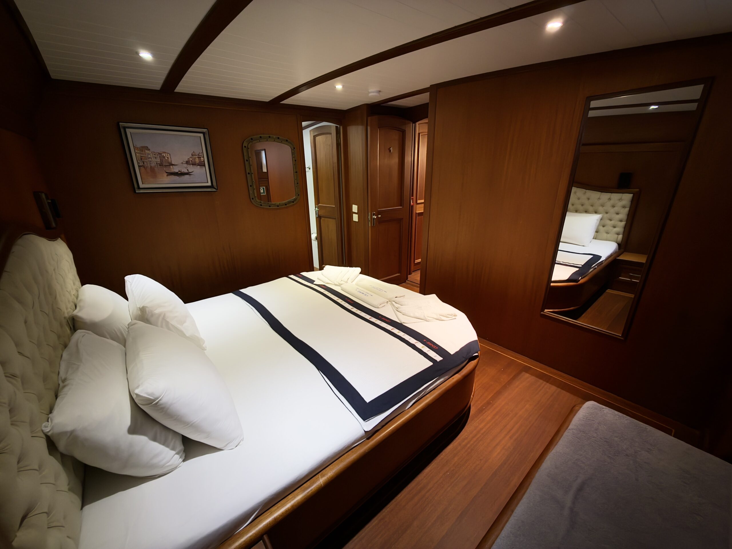 CEMRE 4 A cozy cabin with a double bed, white bedding, and multiple pillows. The room features polished wood walls, a large mirror, a painting, and a doorway leading to another part of the cabin.