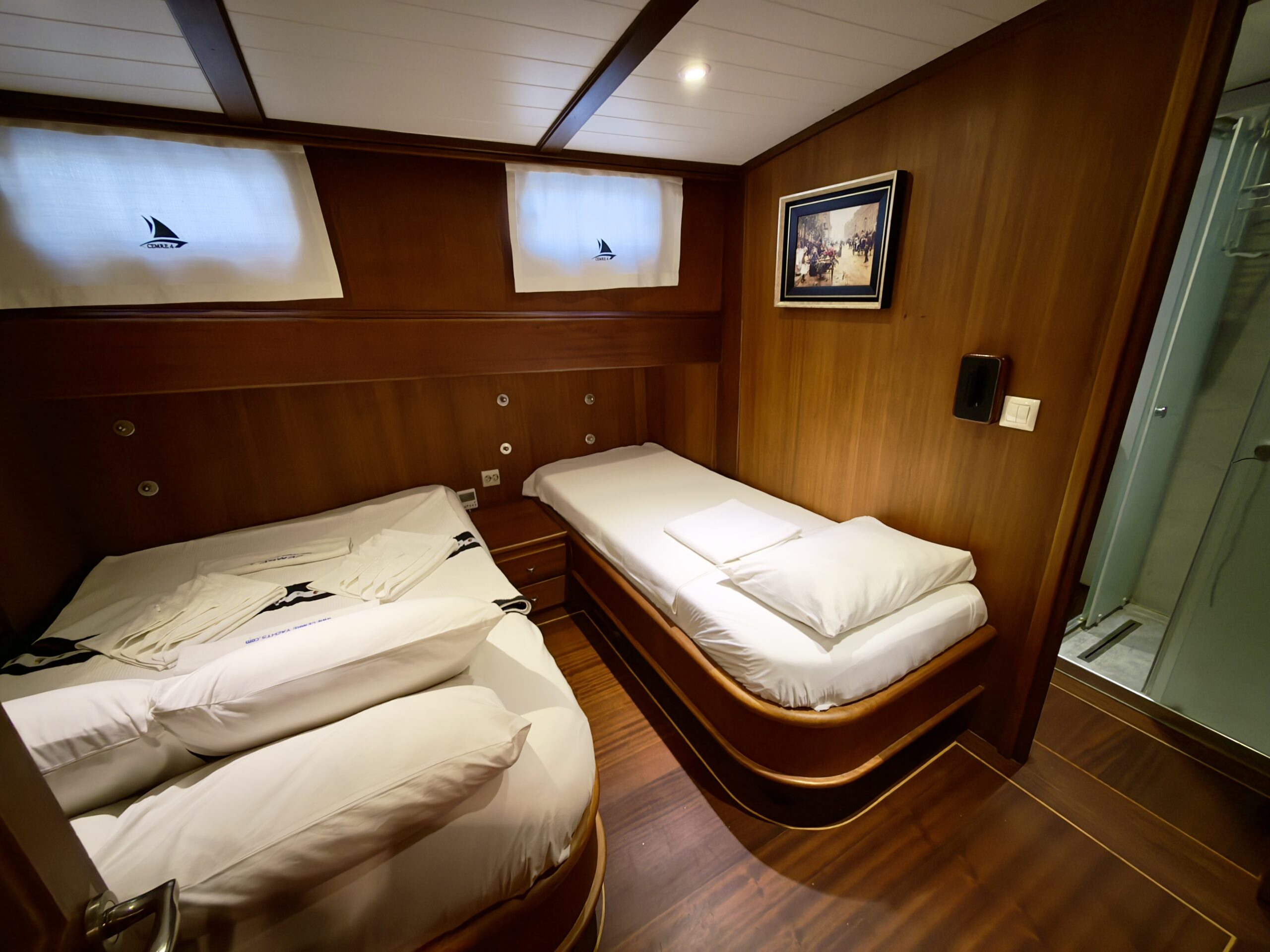 CEMRE 4 A cozy cabin with wood-paneled walls, featuring a double bed and a single bed with white bedding, a framed picture on the wall, and a small bathroom visible through a glass door.