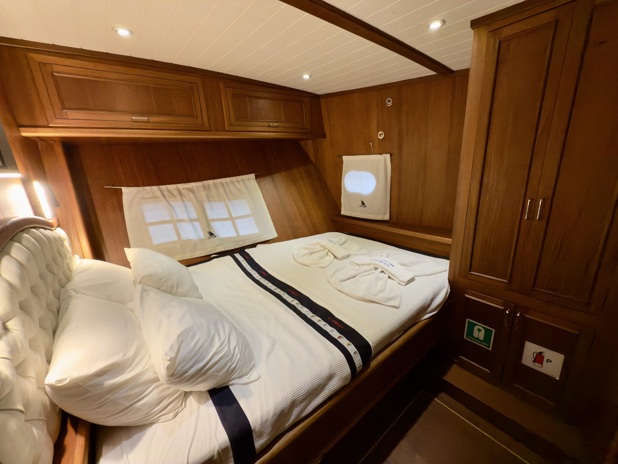 CEMRE 4 A cozy wooden cabin bedroom with a neatly made double bed, white linens, rolled towels shaped like swans, pillows, a small window with curtains, and a wooden wardrobe on the right. Warm lighting creates a relaxing atmosphere.