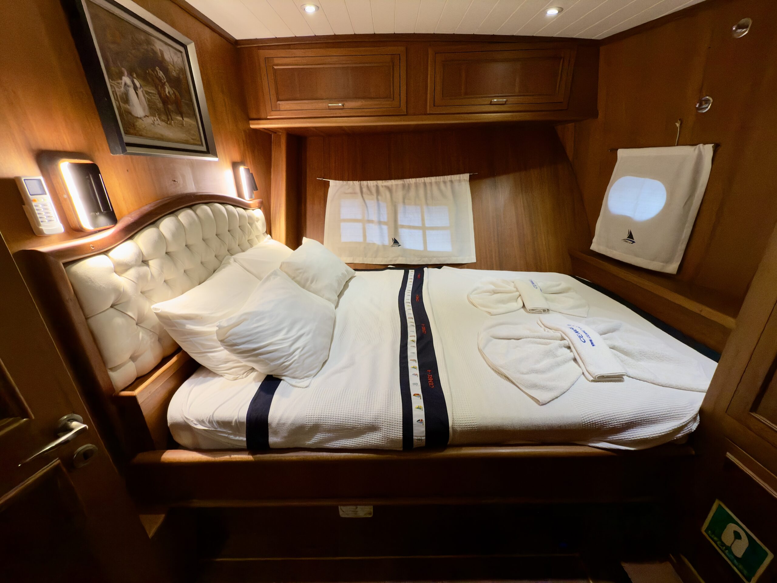 CEMRE 4 A cozy bedroom with wooden walls, a double bed with white bedding, two pillows, towel swans, wall lights, a painting, and small windows with white curtains.