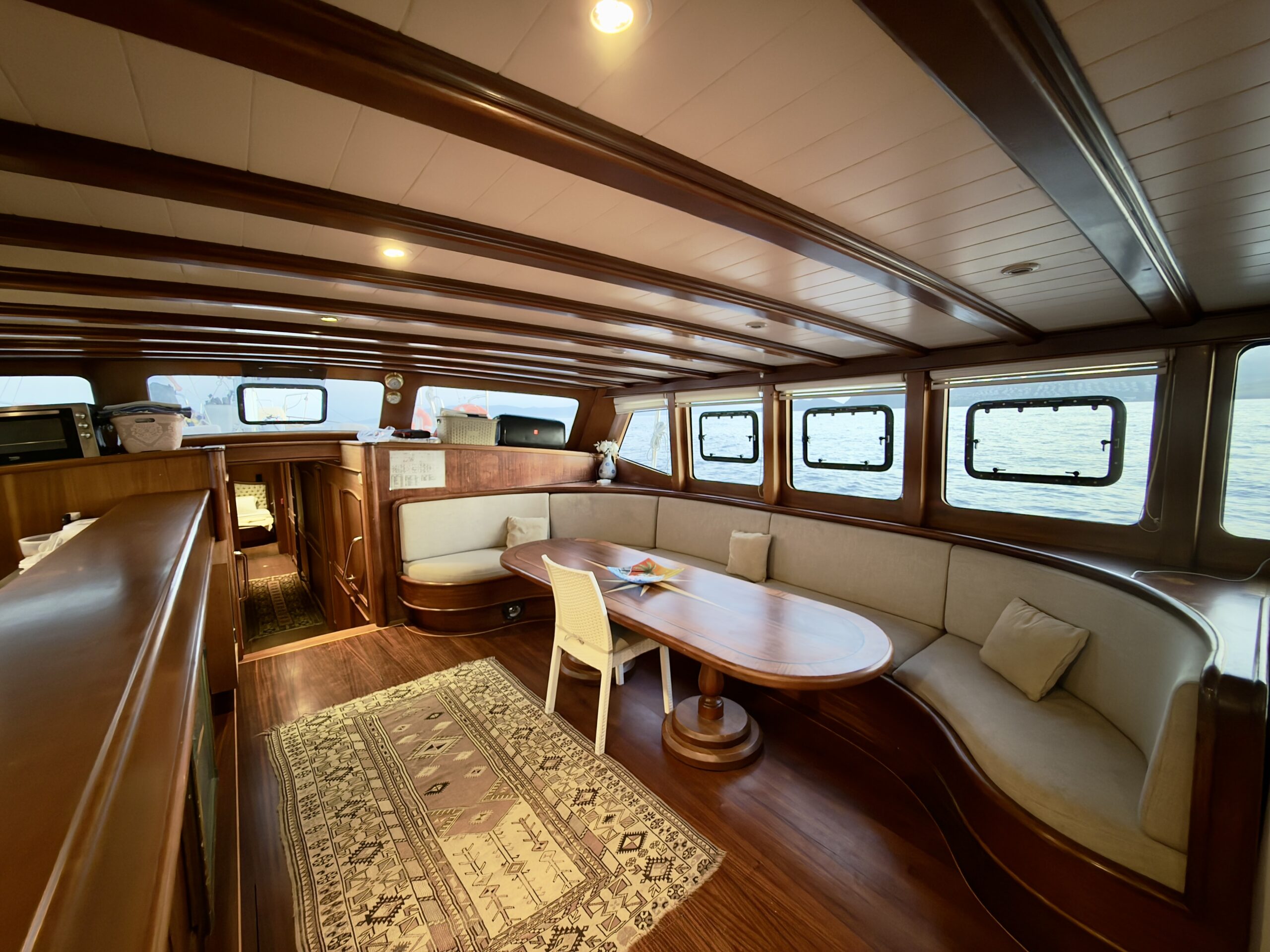 CEMRE 4 Interior of a wooden boat with curved cream sofas, a wooden table, a white chair, and a patterned rug. Windows offer a view of the water outside. Warm lighting creates a cozy atmosphere.