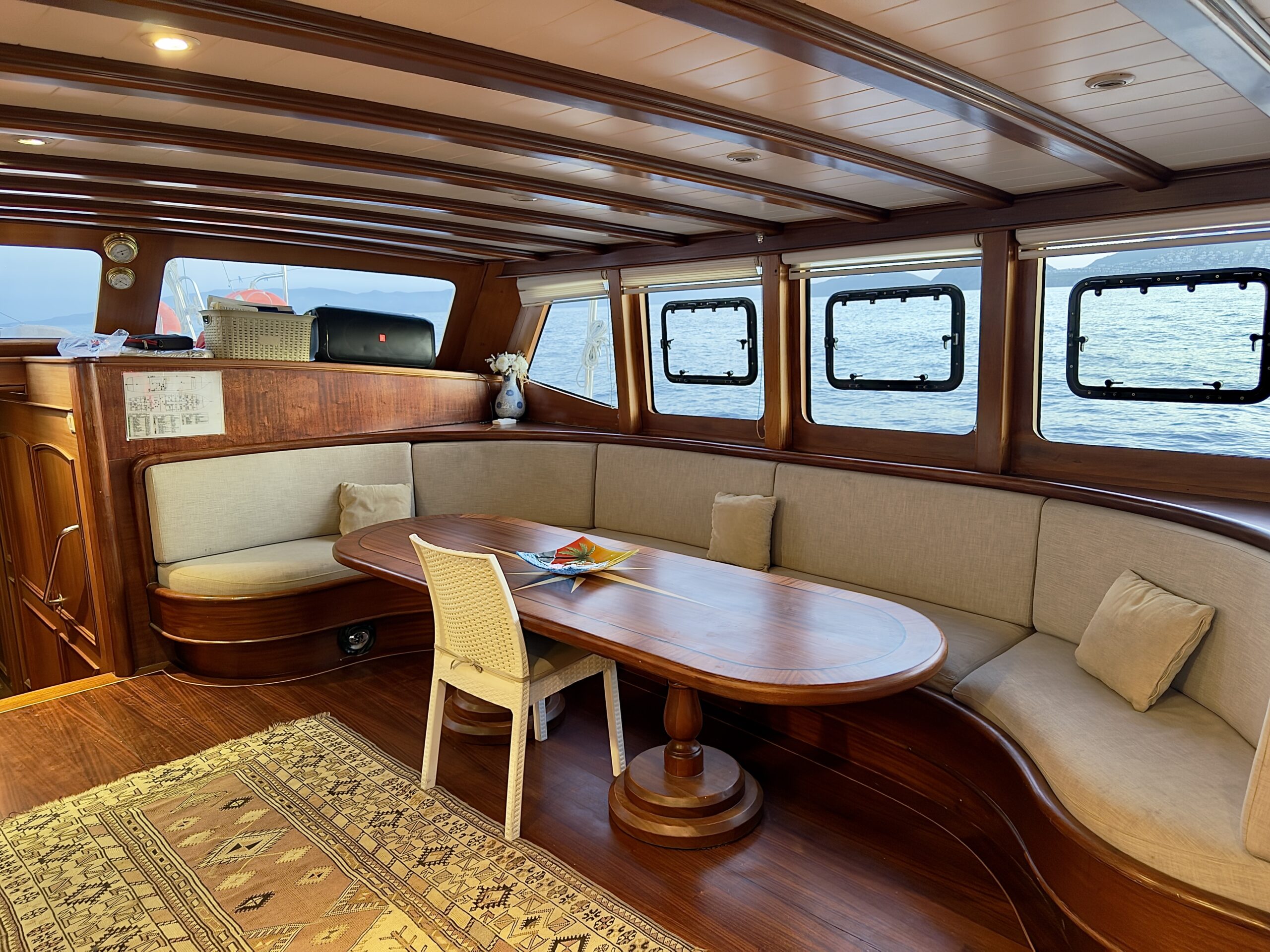 CEMRE 4 A cozy yacht interior featuring a curved beige sofa, a wooden dining table with a white chair, large windows overlooking the sea, and a patterned rug on a polished wooden floor.