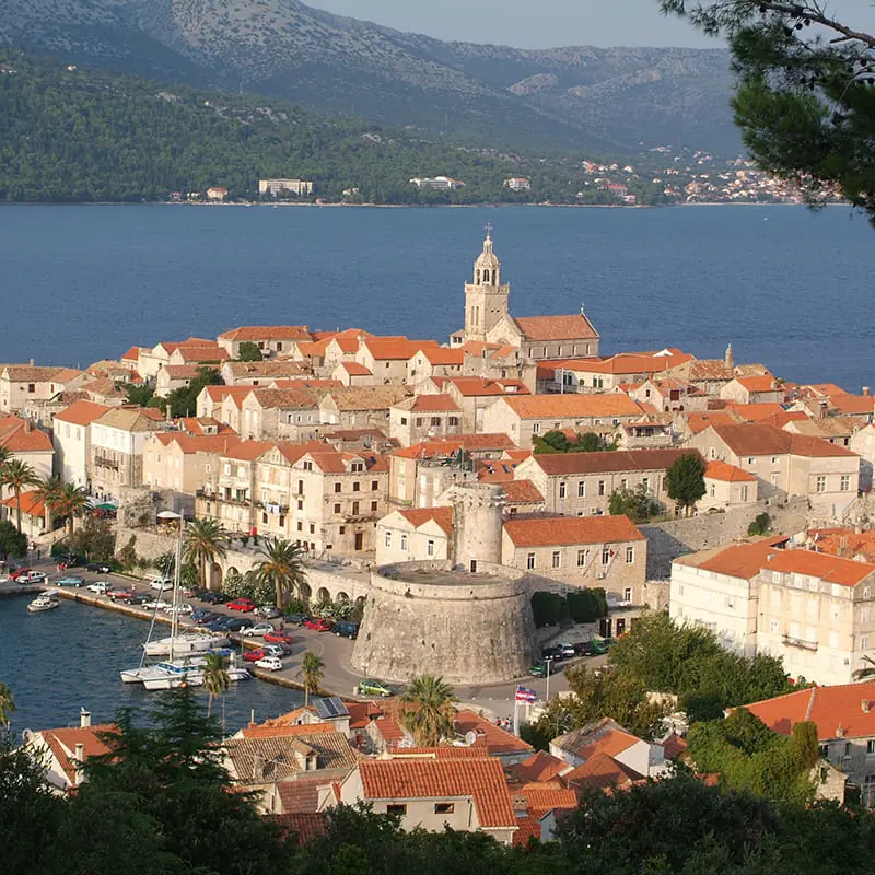 A coastal town with red-roofed buildings, a prominent stone church tower, a round fort, a marina with boats, and mountains in the background under a clear sky.
