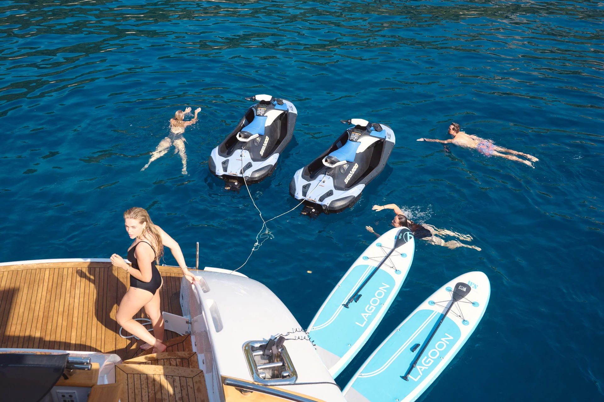 A woman steps off a boat into the blue sea where two jet skis, two paddleboards, and three other people are floating.