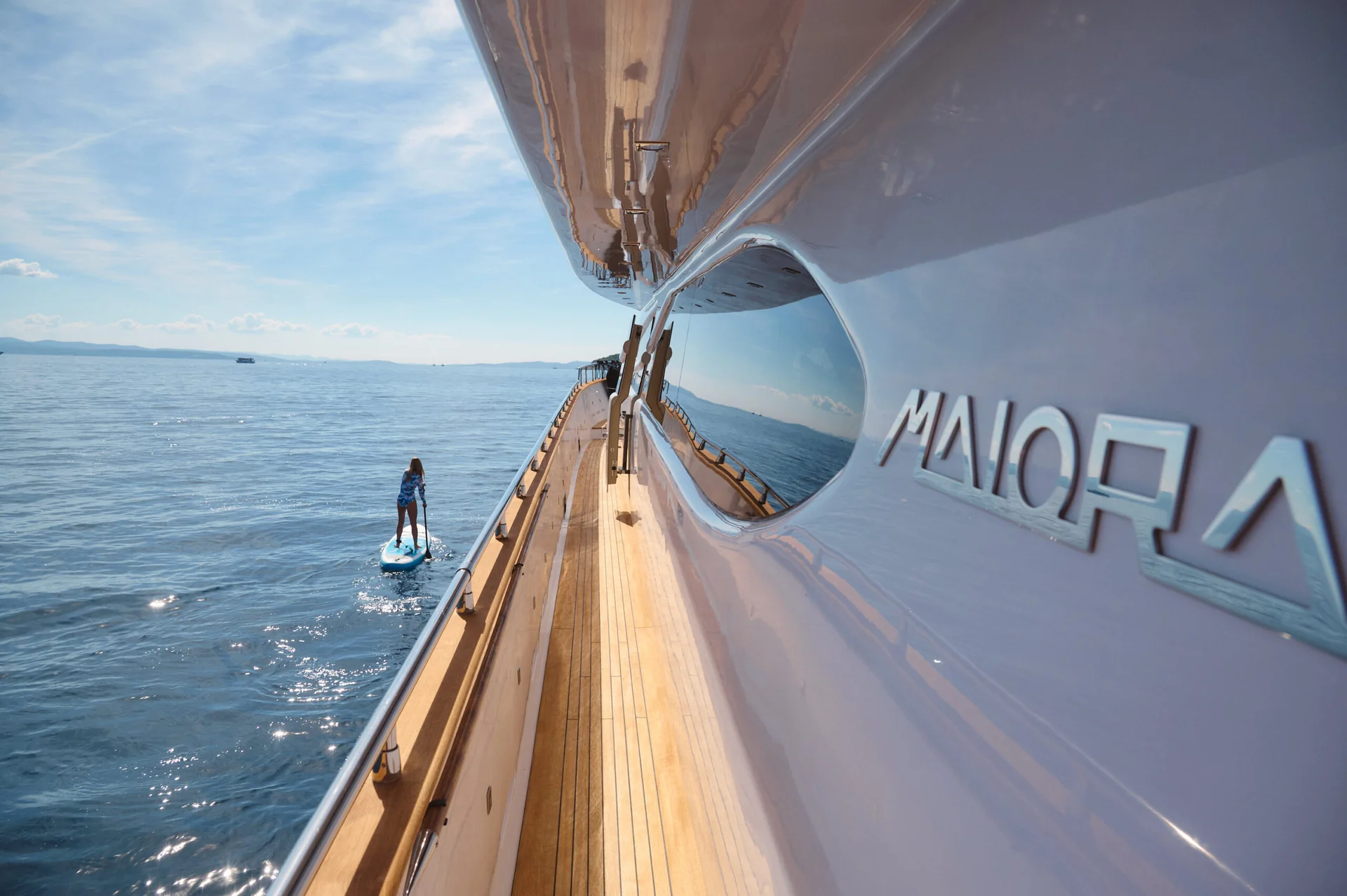 A person paddleboarding on the ocean alongside the teak deck of a Maiora yacht.