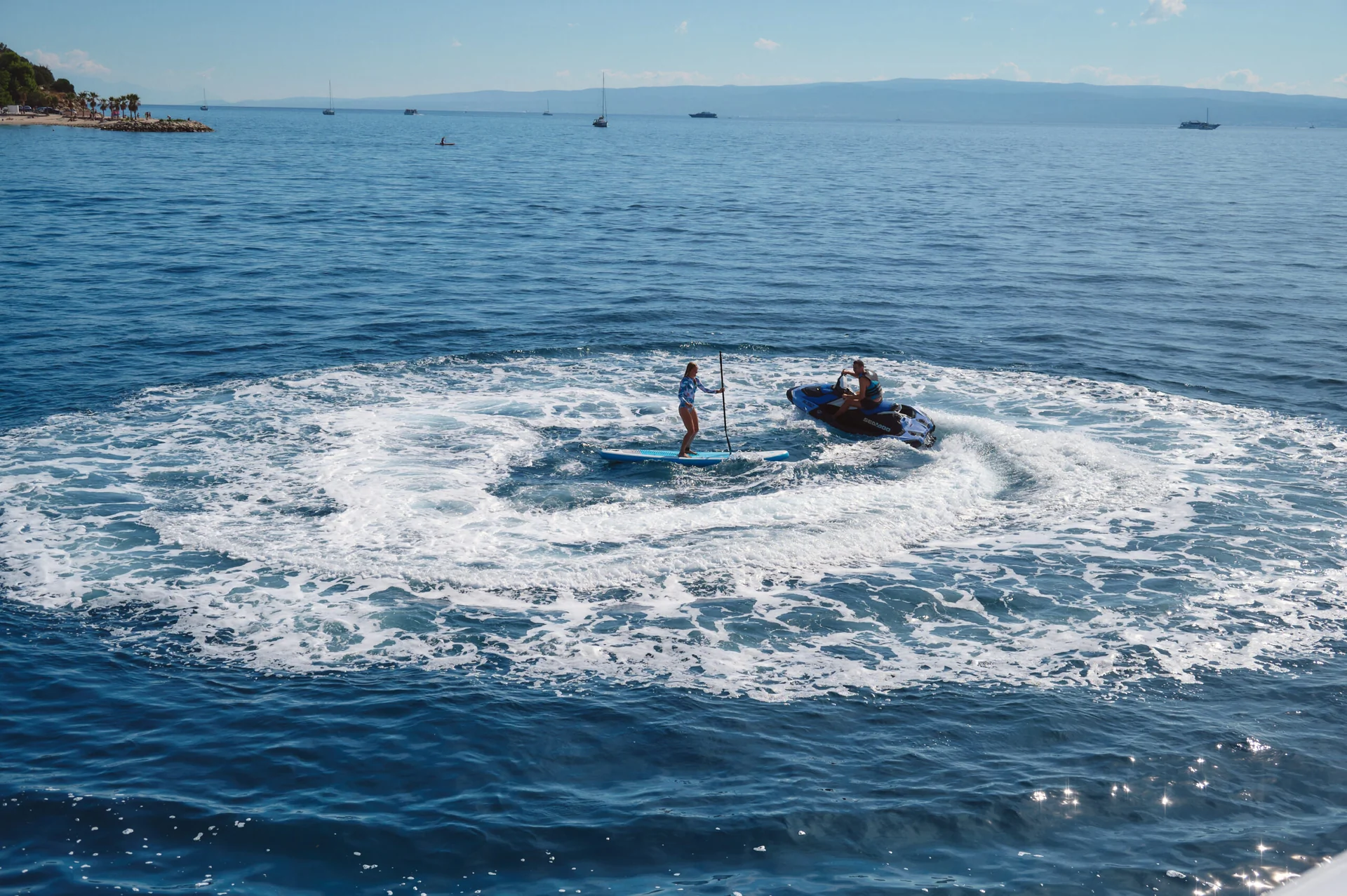 A person on a stand-up paddleboard stands in the center of a circular wake created by a jet ski circling them on the ocean.