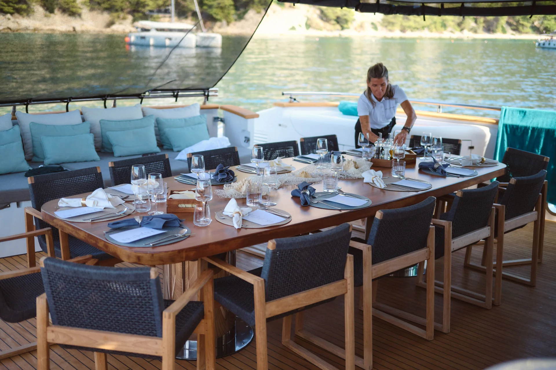 A crew member sets a wooden dining table on the deck of a yacht, with place settings prepared for a meal.