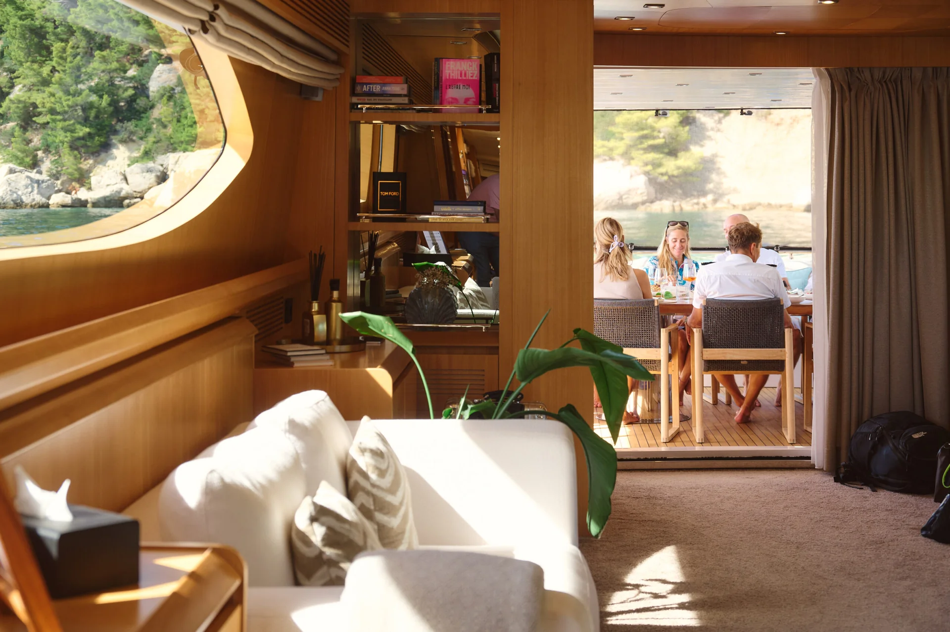 Interior of a yacht with a white sofa in the foreground and a group of people dining on the outdoor deck.