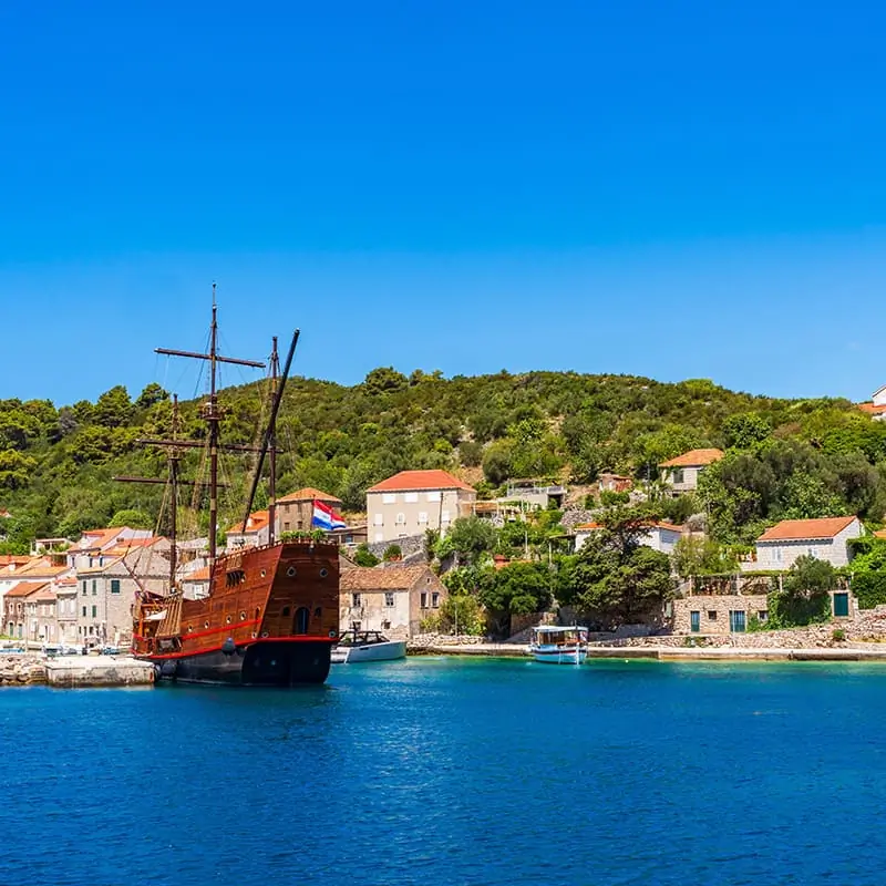 A wooden sailing ship docked by a small coastal village with red-roofed houses, lush green hills, and clear blue water under a bright, sunny sky.