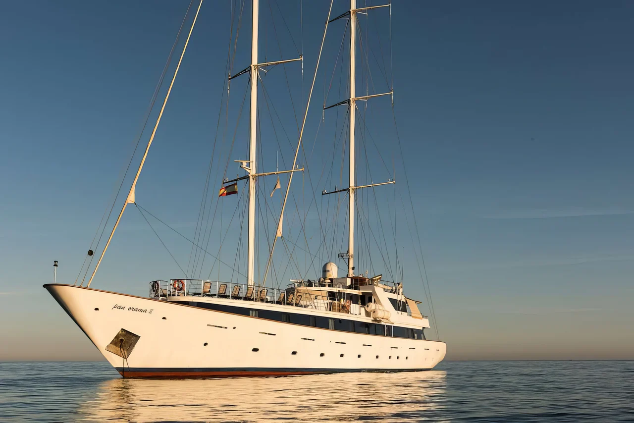 A large white sailing yacht with three tall masts floats on calm water under a clear blue sky, with sunlight reflecting off the surface.