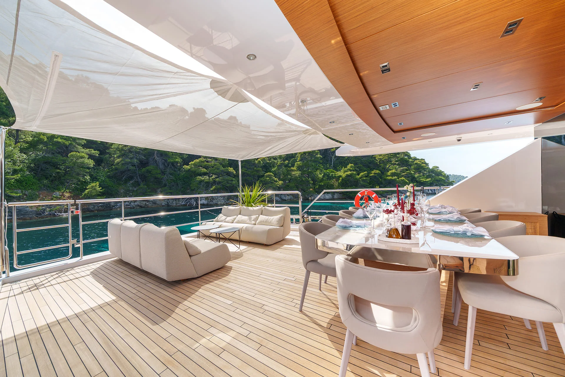 A luxury yacht deck features a shaded dining table set for a meal, cushioned lounge chairs, wooden flooring, and a view of trees and calm water in the background.