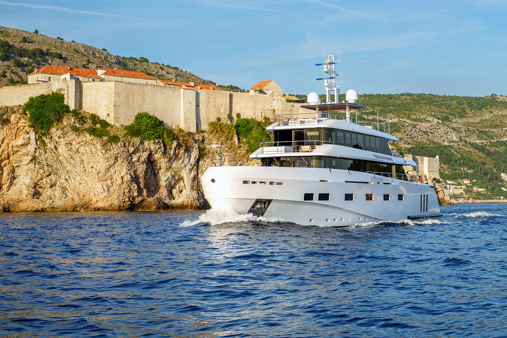 A white luxury yacht cruises on blue water near rocky cliffs with historic stone buildings and green hills in the background under a clear sky.