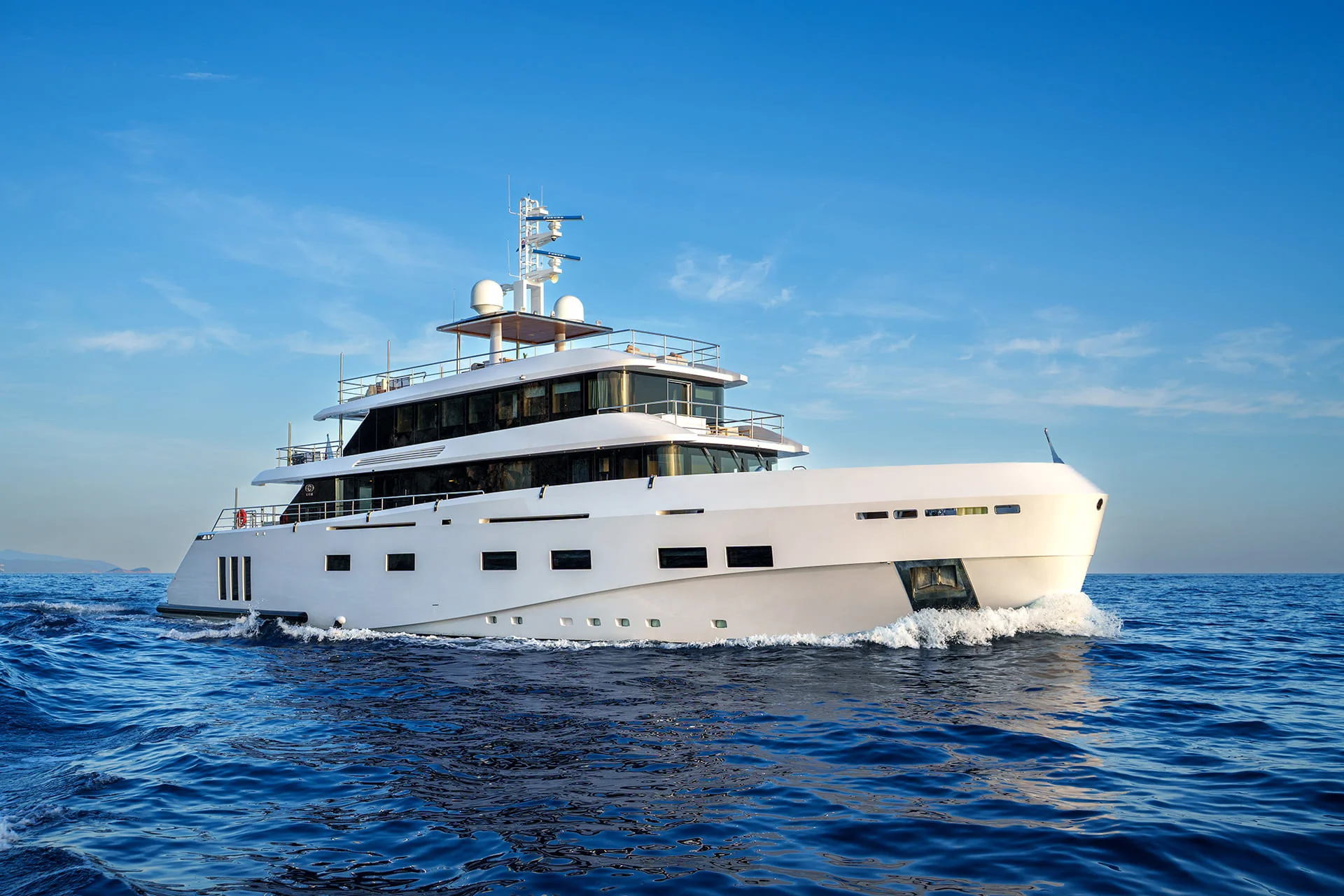 A large white luxury yacht sails on calm blue ocean water under a clear sky, creating small waves at the bow.
