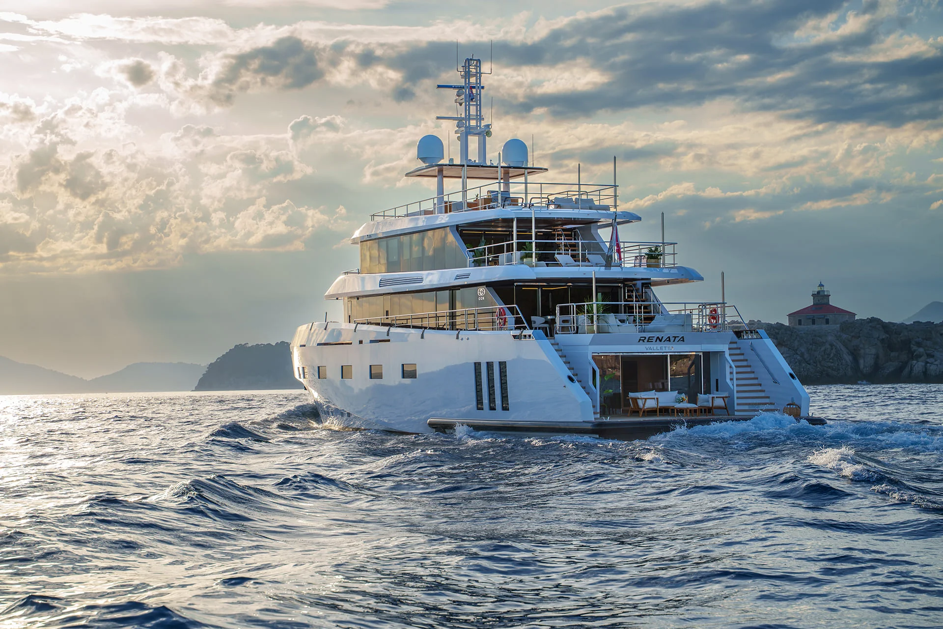 A large white yacht named RENATA cruises on blue water near a coastline, with a small lighthouse and rocky hills in the background under a cloudy sky at sunset.