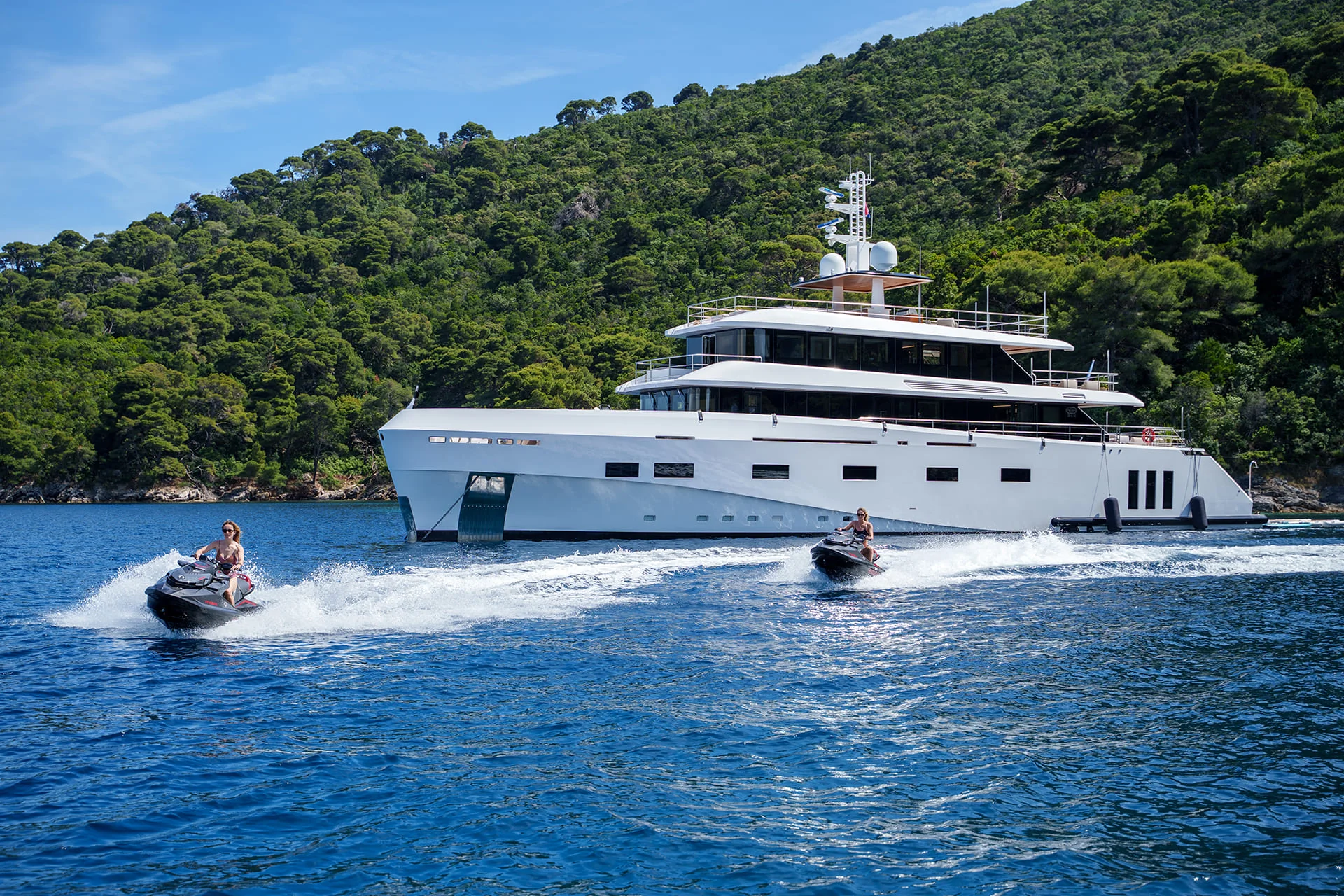 A large white yacht is anchored near a lush green hillside. Two people ride jet skis on the blue water in front of the yacht, creating small waves as they speed by. The sky is clear and sunny.