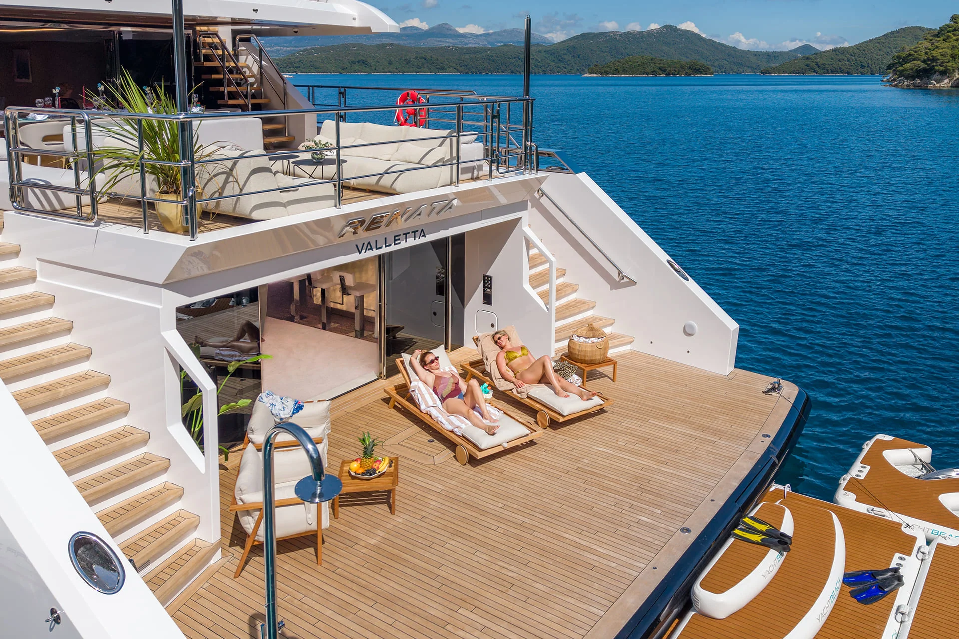 Two people relax on lounge chairs on the deck of a luxury yacht, docked in clear blue water with green islands in the background. The scene is sunny and peaceful, with steps leading up and down from the main deck.