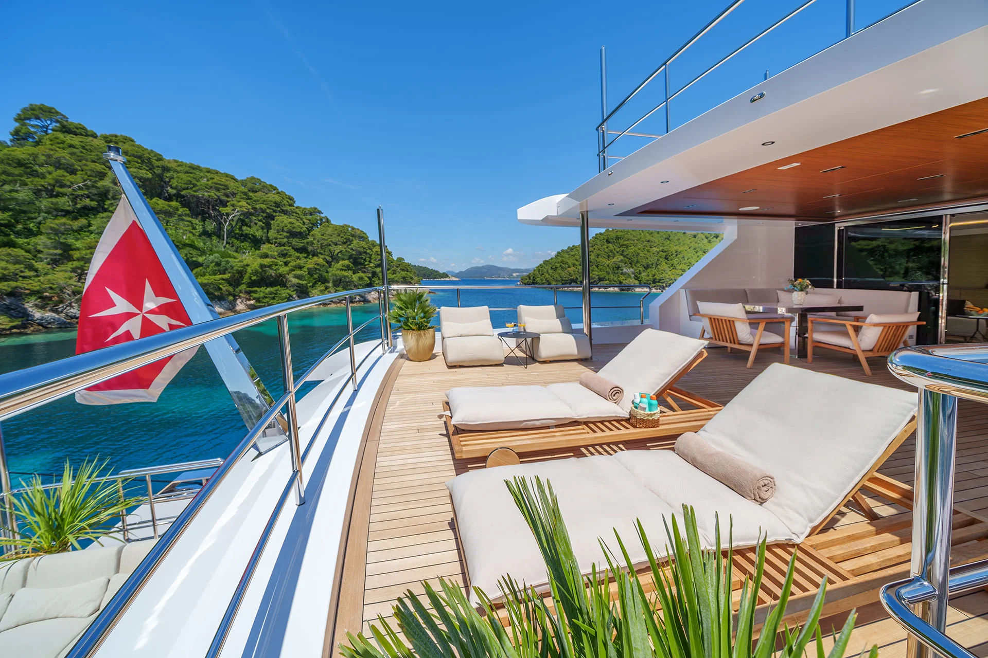 Sunny yacht deck with cushioned lounge chairs, potted plants, a table, and a red flag. The yacht is docked near green islands on clear blue water, with a bright blue sky overhead.