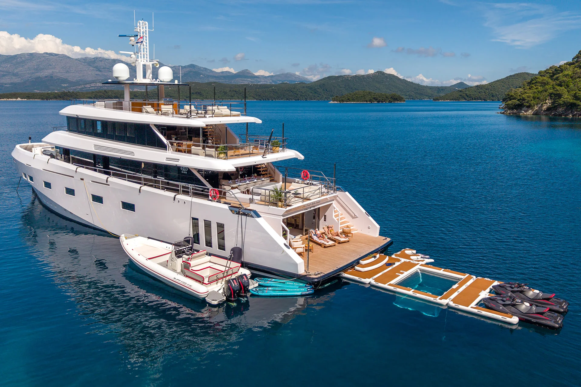 A large white yacht anchored in calm blue water with mountains in the background. Inflatable docks, jet skis, paddleboards, and a small pool float beside the yacht.