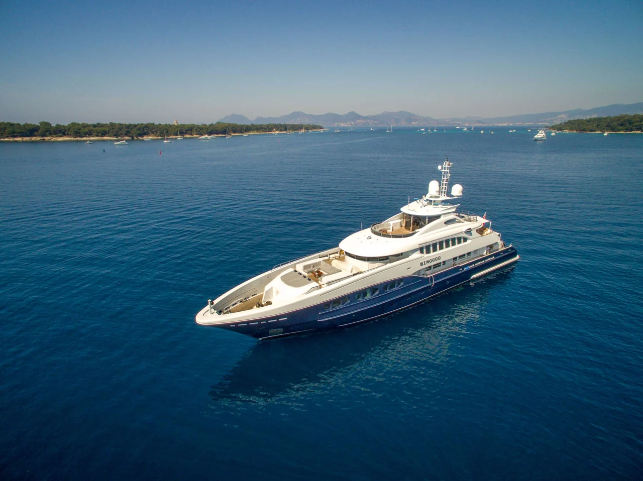 A large luxury yacht is anchored in calm blue water near a coastline with trees and distant mountains under a clear sky.