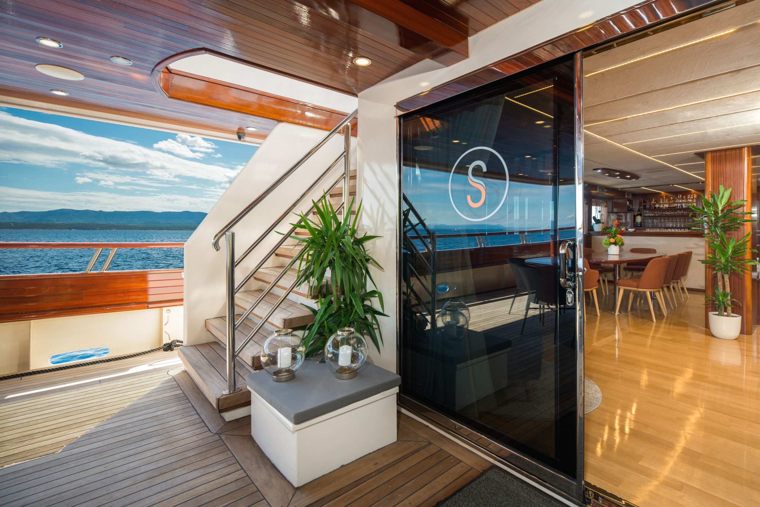 SON DE MAR Luxury yacht deck with wood accents, potted plants, and glass doors leading to a modern interior lounge and dining area. Scenic ocean and distant coastline visible under a partly cloudy sky.