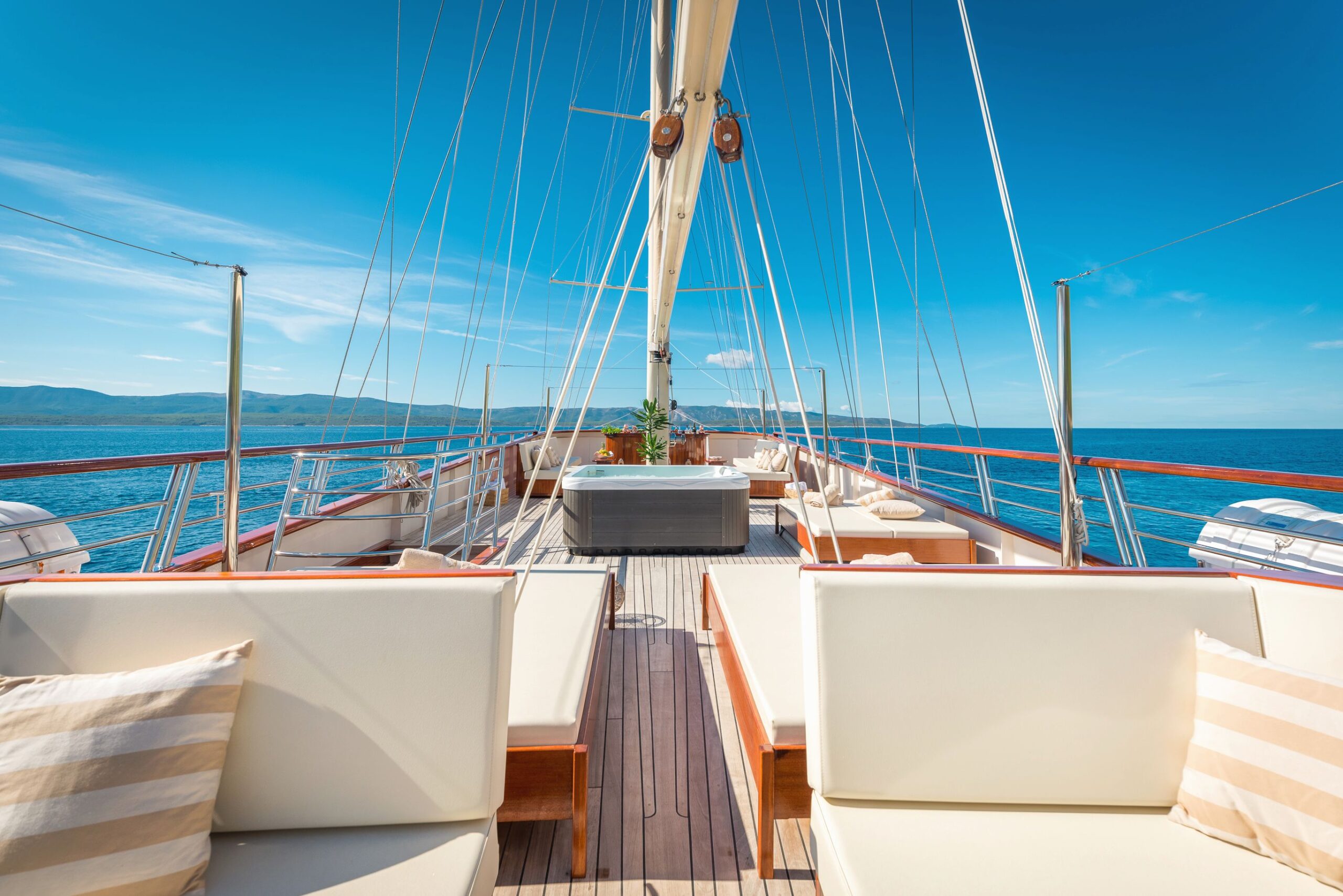SON DE MAR Sunny deck of a luxury yacht with cushioned seating, striped pillows, and a hot tub, overlooking calm blue sea and distant coastline under a clear sky.