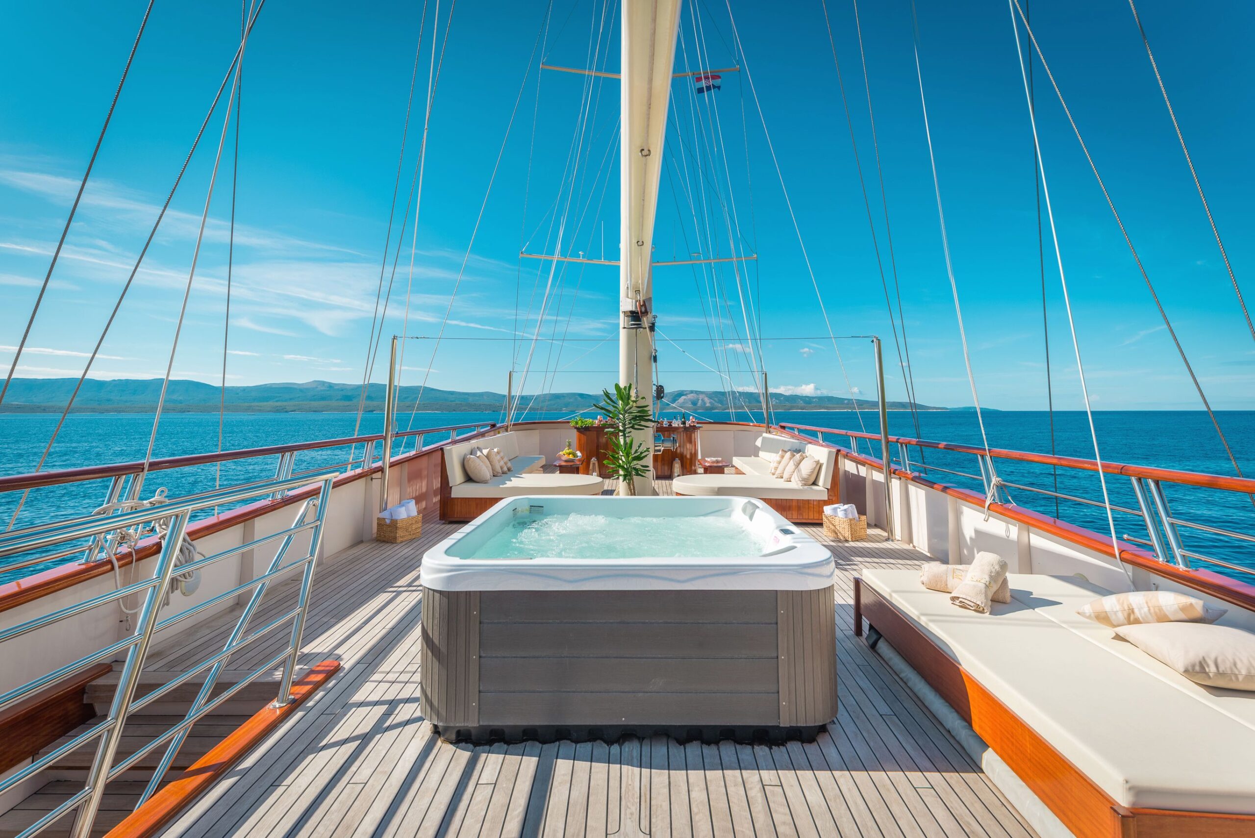 SON DE MAR A luxury yacht deck with a hot tub, sun loungers, and cushions, overlooking calm blue sea and distant green hills under a bright, clear sky.