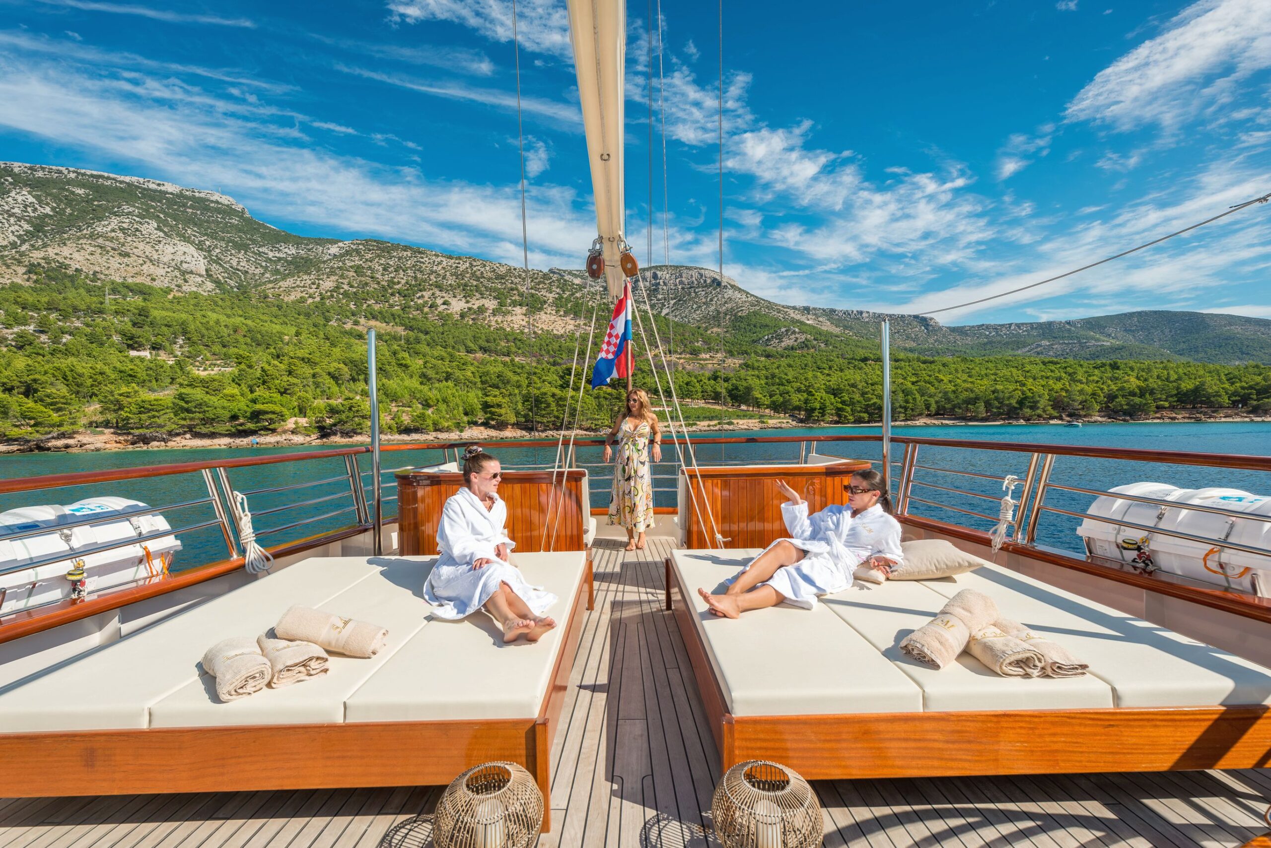 SON DE MAR Two people in white robes relax on sunbeds on the deck of a yacht, surrounded by blue water and green hills under a bright, clear sky. A third person stands at the center of the deck.