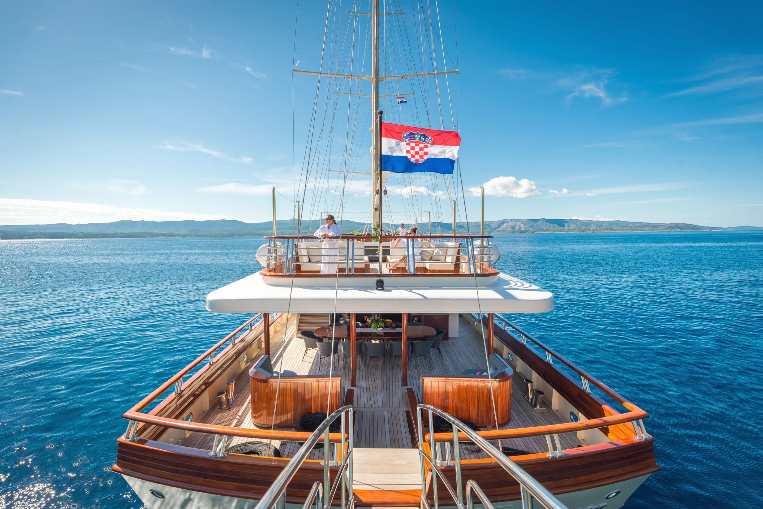SON DE MAR A yacht with a Croatian flag sails on bright blue water under a clear sky. Two people stand on the upper deck, and distant mountains are visible along the horizon.
