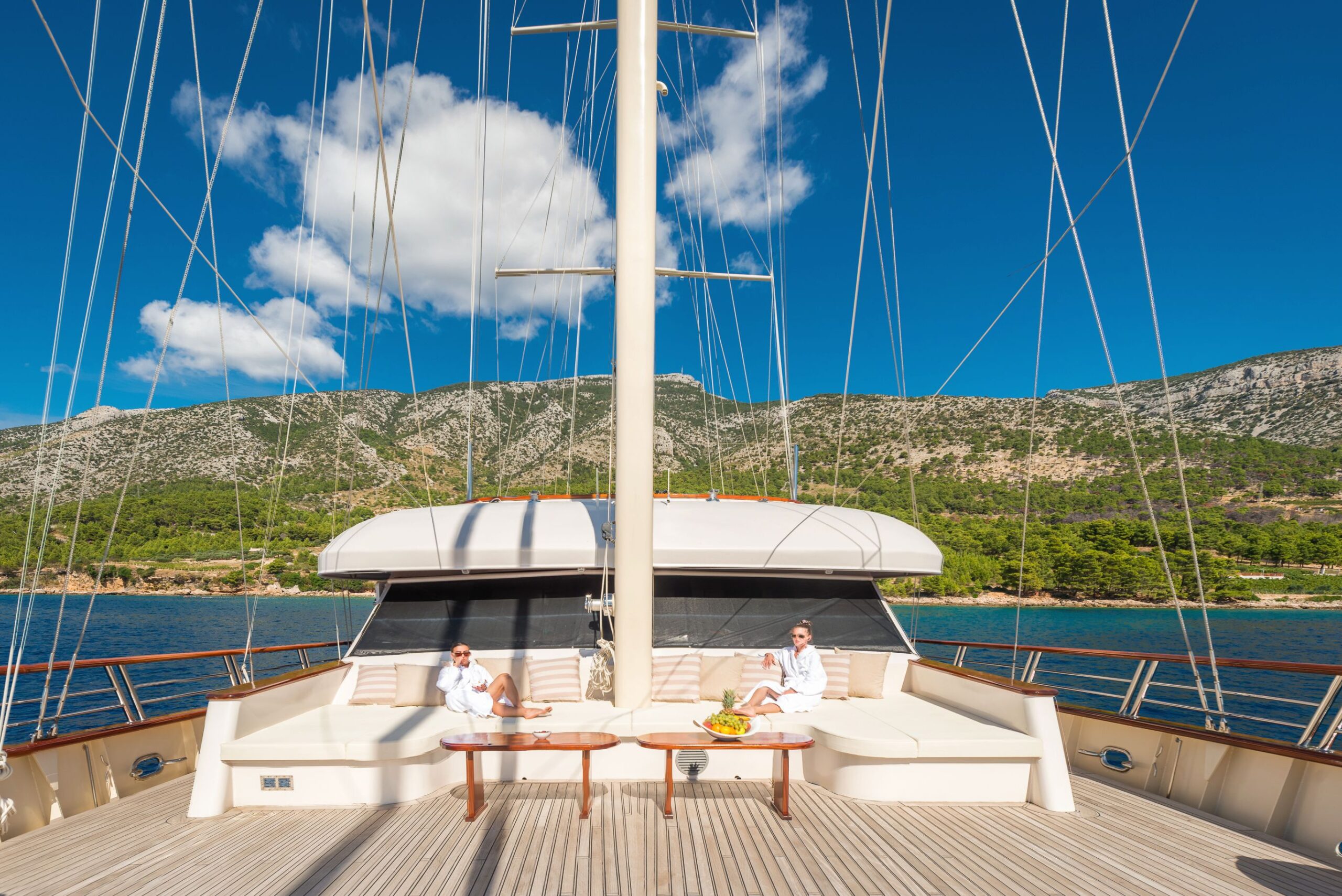 SON DE MAR Two people in white clothing relax on the deck of a luxury yacht, sitting on cushioned benches around a table with fruit. The yacht is near a coastline with green hills and a clear blue sky.