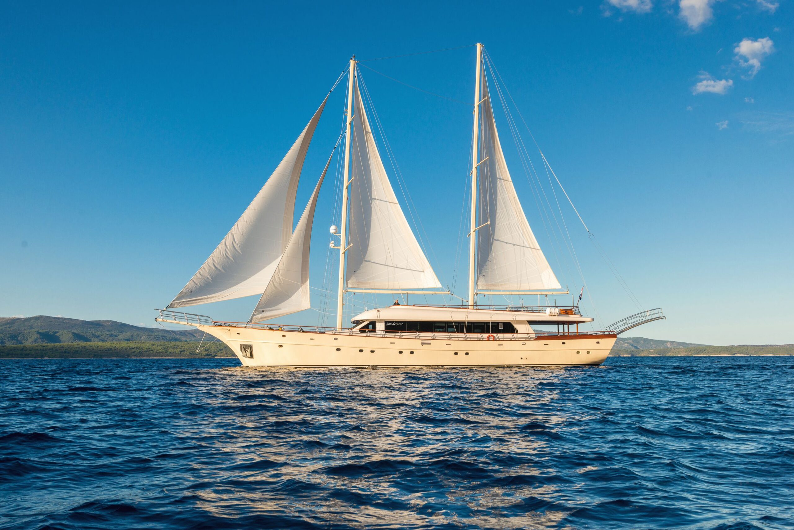 SON DE MAR A large sailboat with two masts and white sails floats on deep blue water under a clear sky, with green hills visible in the background.
