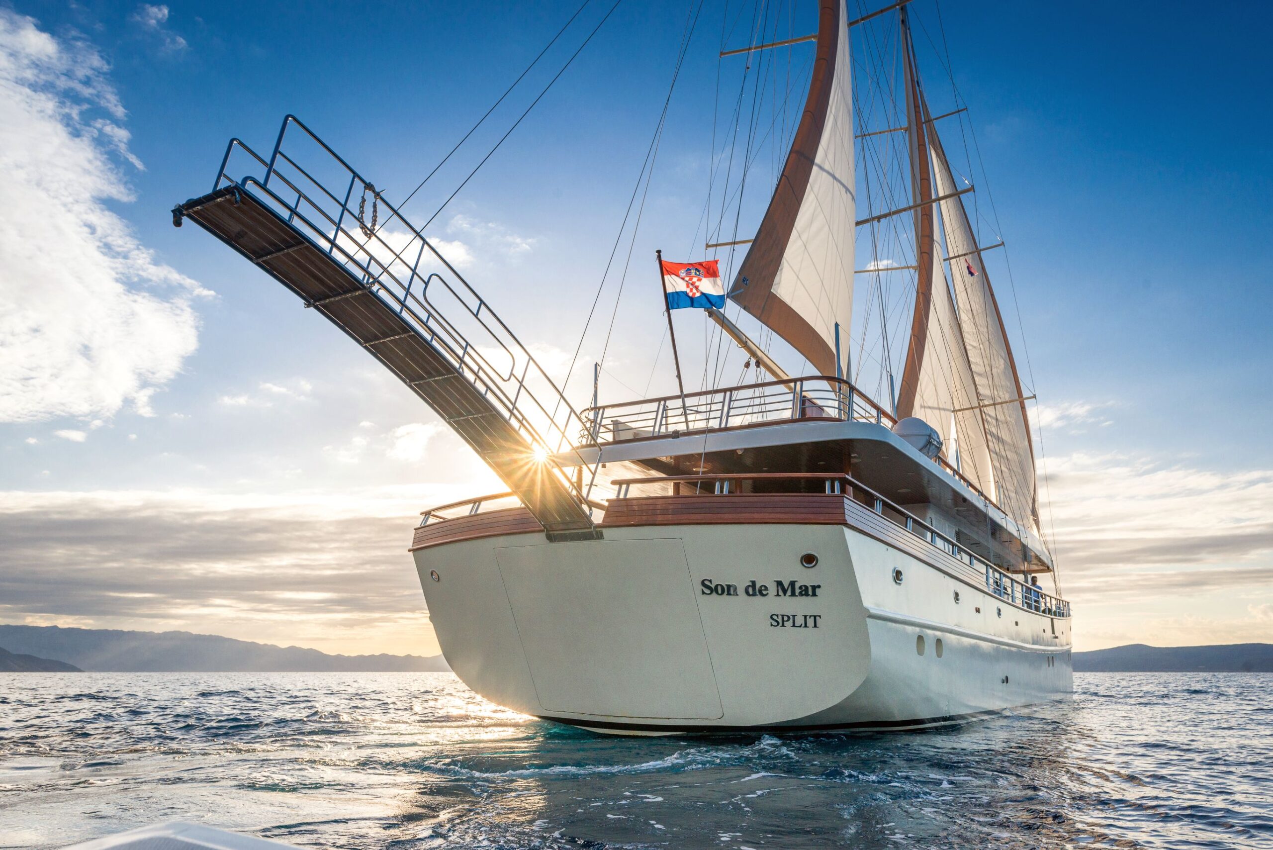 SON DE MAR A large white sailing yacht named “Son de Mar” with a raised gangway sails on the ocean at sunset, flying the Croatian flag, with mountains and a partly cloudy sky in the background.