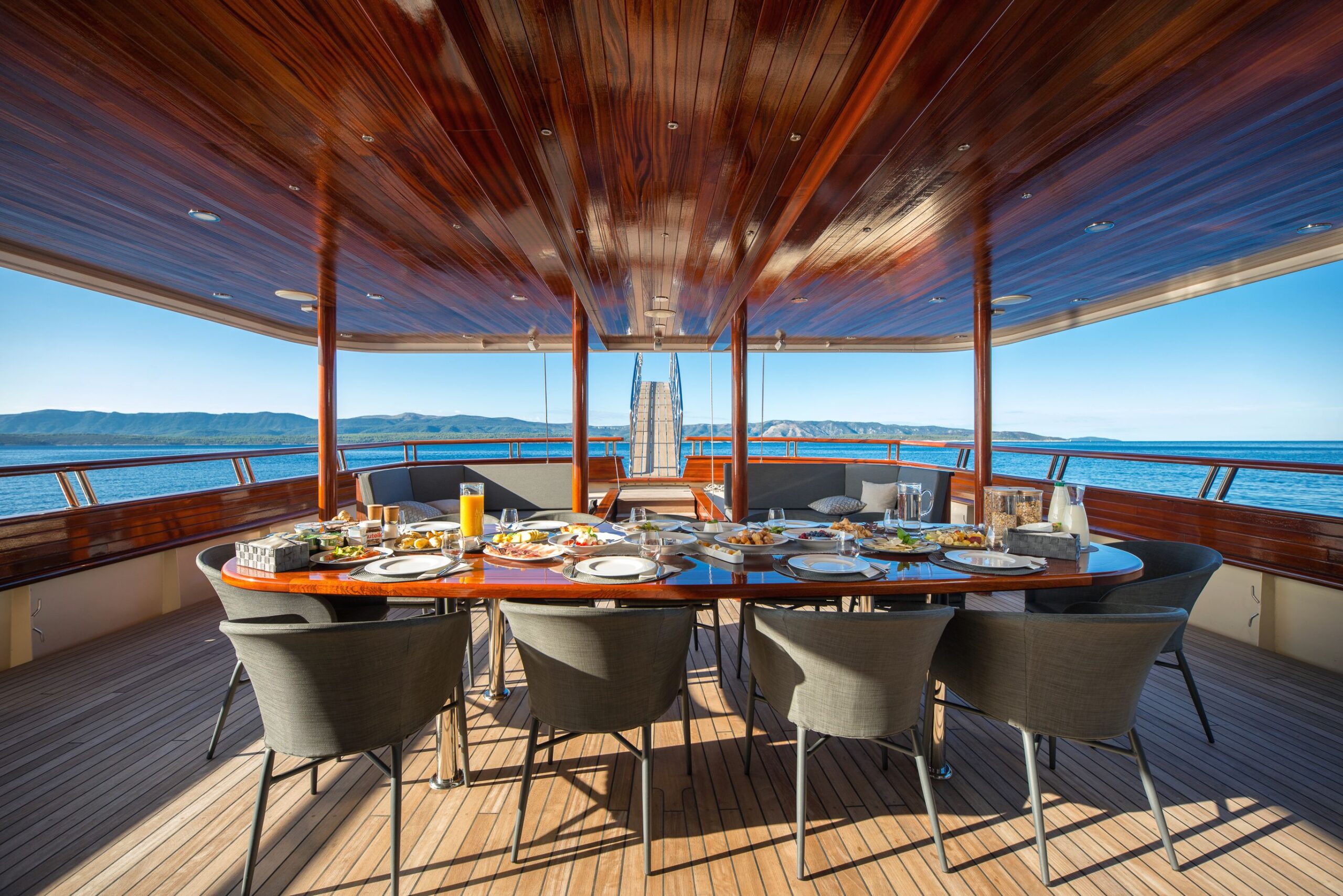 SON DE MAR A luxurious yacht dining area with a large oval table set for a meal, surrounded by chairs, overlooking calm blue water and distant mountains under a clear sky.